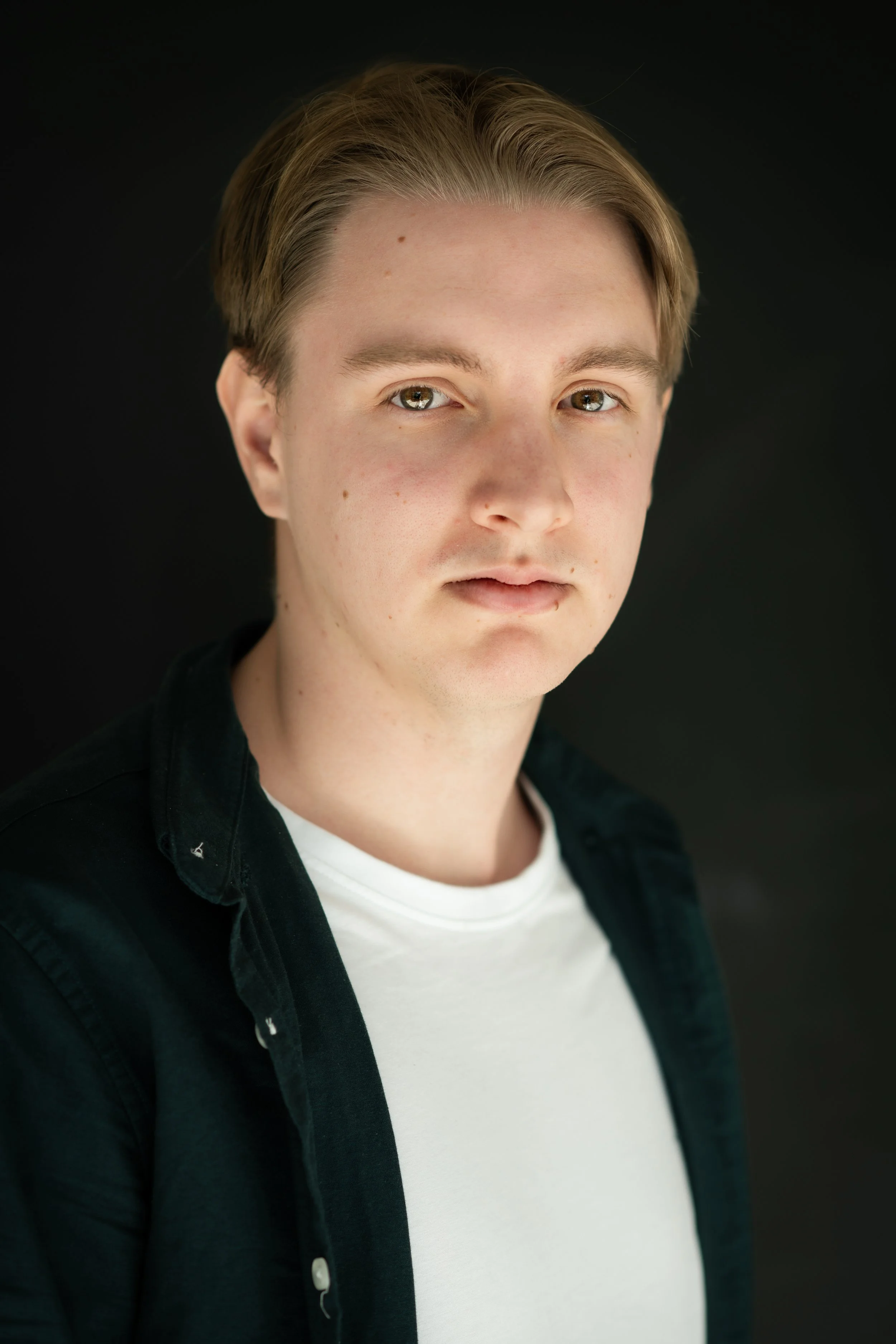 Close-up of a young man with light skin, styled blonde hair, and hazel eyes, wearing a black shirt over a white t-shirt, against a dark background.