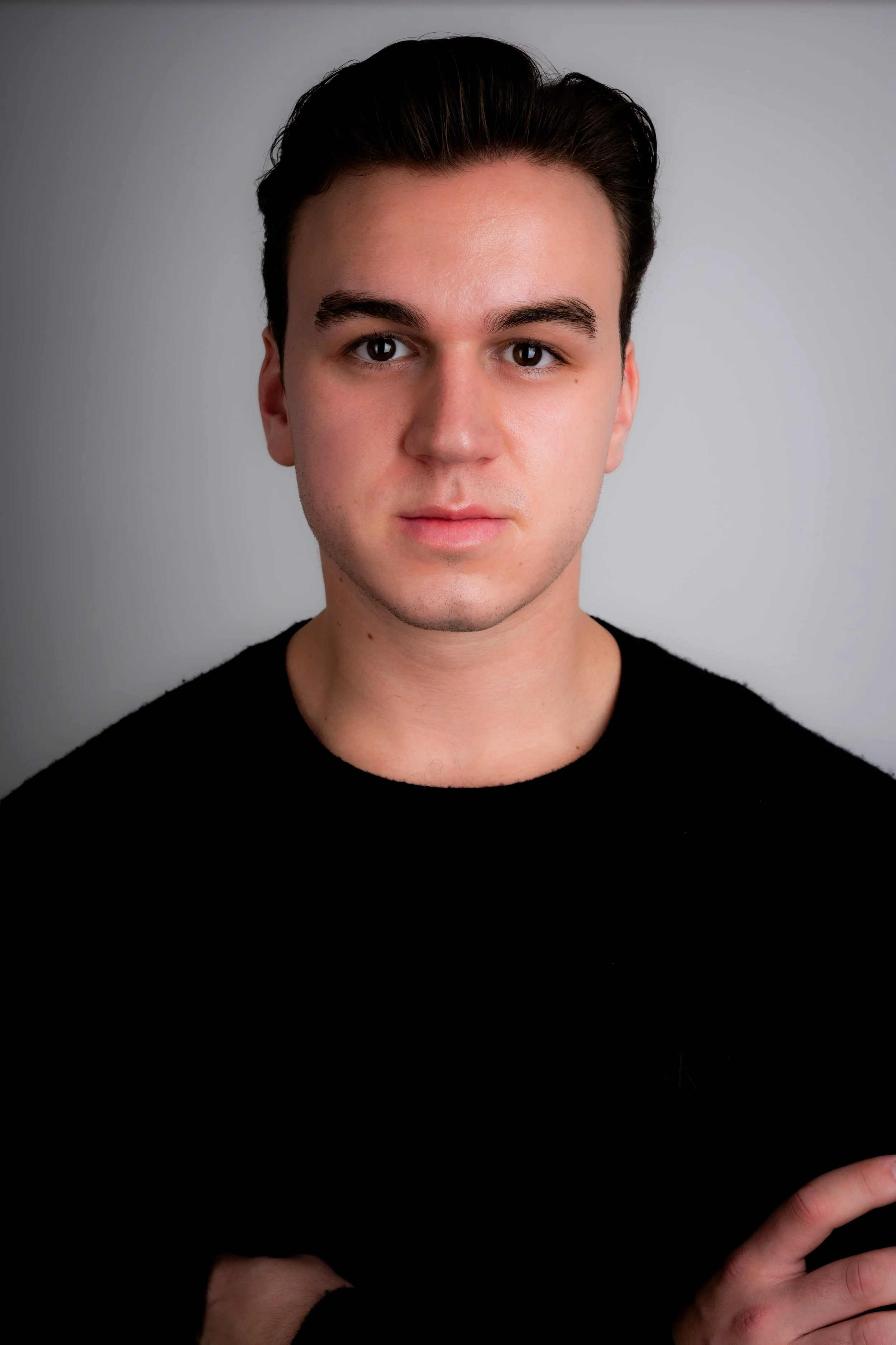 Portrait of a young man with dark hair, wearing a black sweater, standing against a plain gray wall.