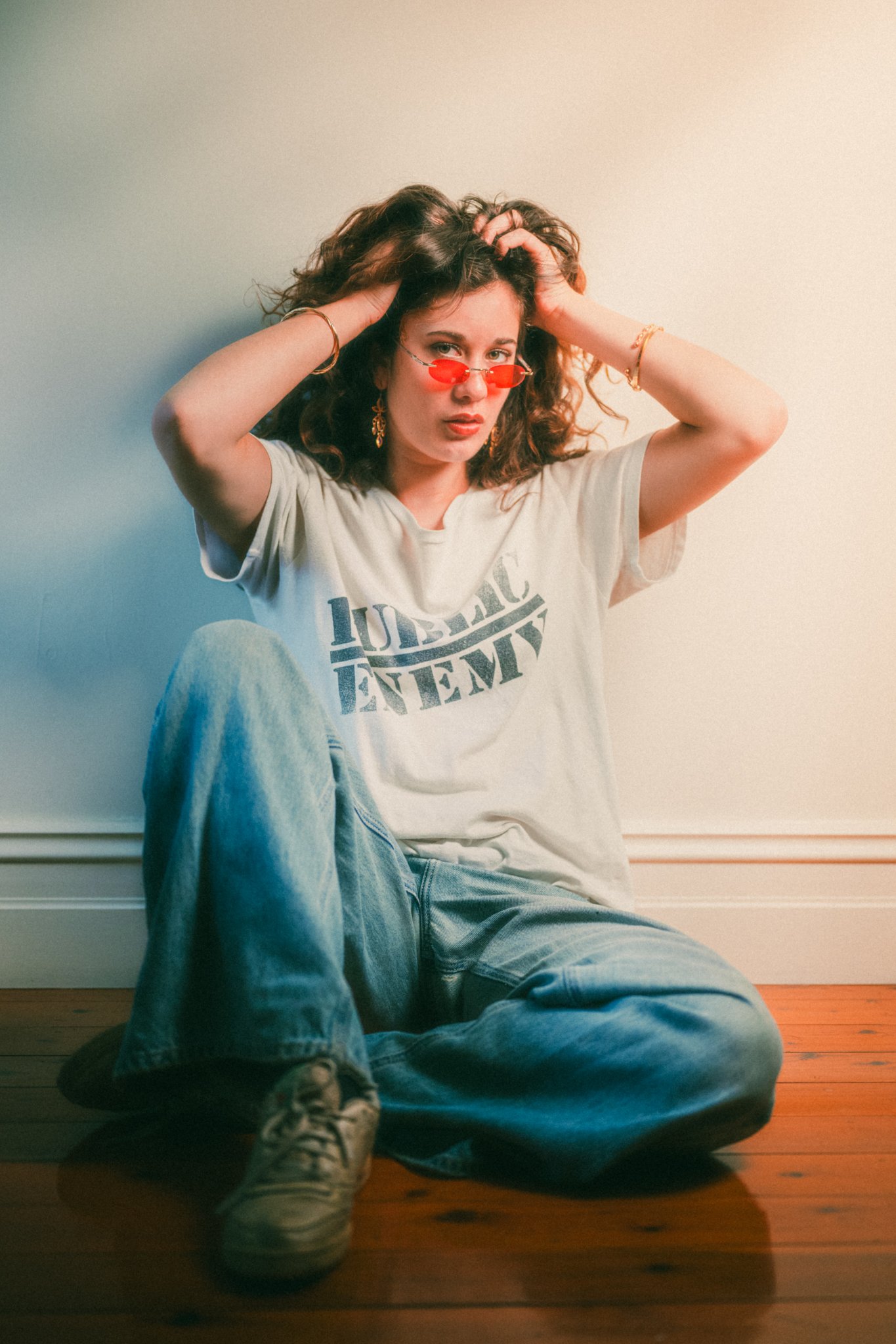 A woman with curly hair wearing sunglasses, a white t-shirt with black text, ripped jeans, and sneakers, sitting on a wooden floor with one leg crossed over the other, posing with her hands in her hair in front of a plain wall.