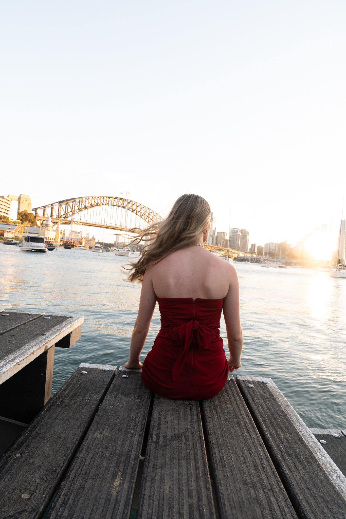 A woman sitting on a wooden dock overlooking a river at sunset, with a city skyline and the Sydney Harbour Bridge in the background.
