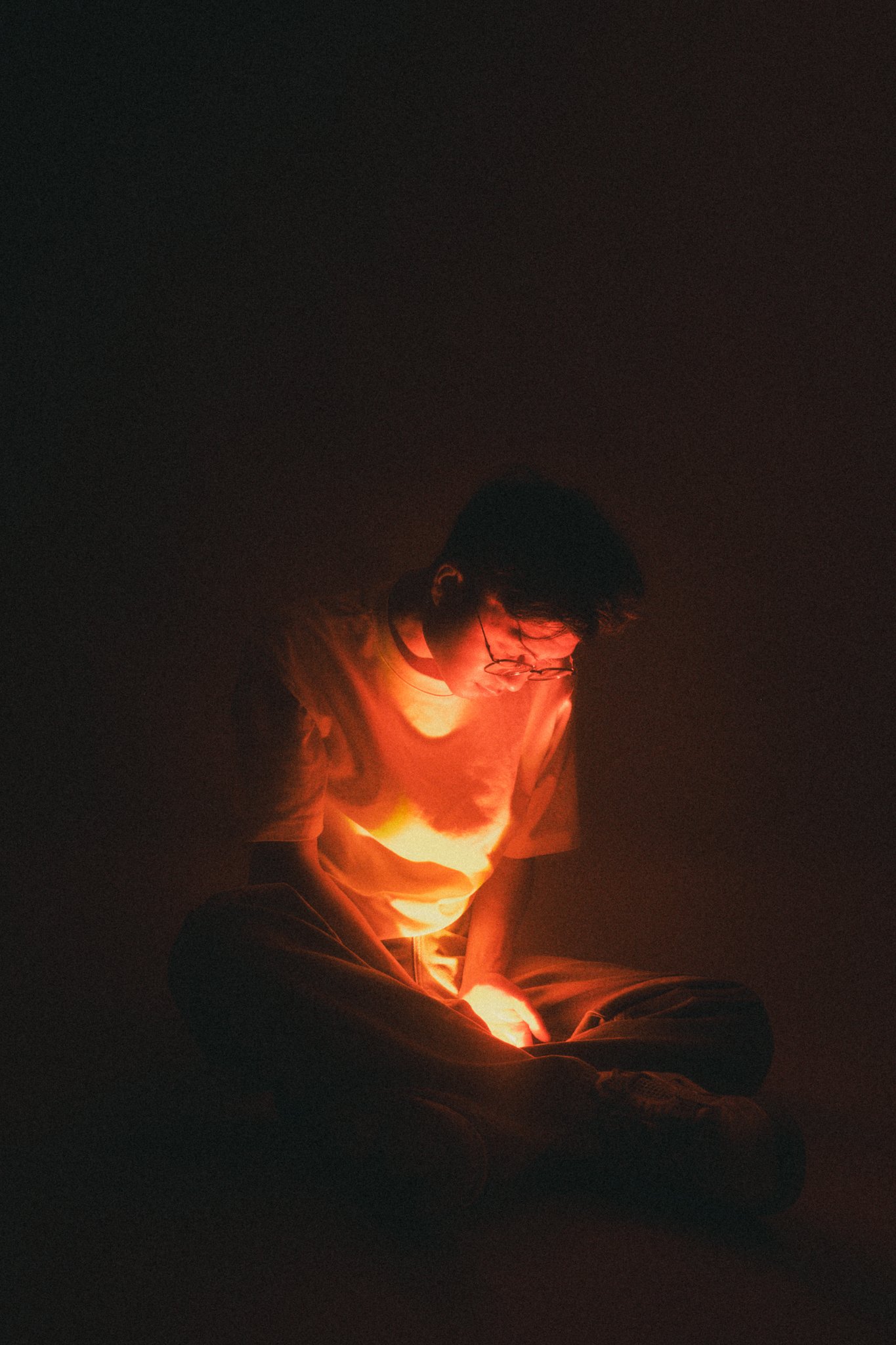 A person wearing glasses sitting cross-legged in a dark room illuminated by a red glow.