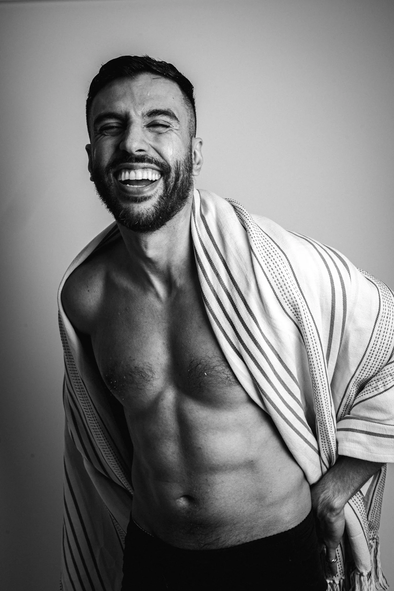 A smiling man with a beard and short hair, shirtless with a striped towel draped over his shoulder, standing against a plain background.