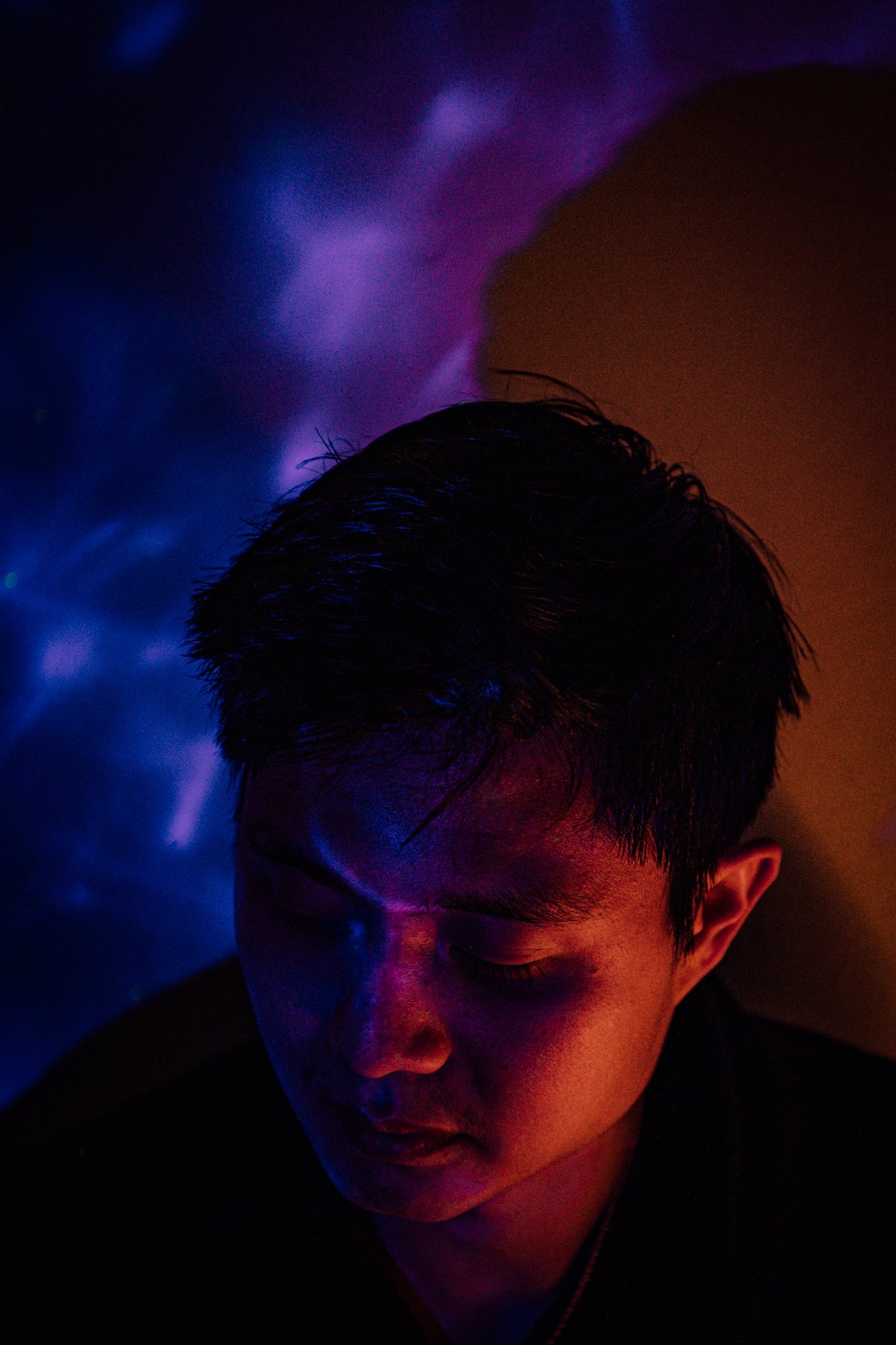 A young man with dark hair, resting with eyes closed, illuminated by colorful neon lights in purple, blue, and orange hues.