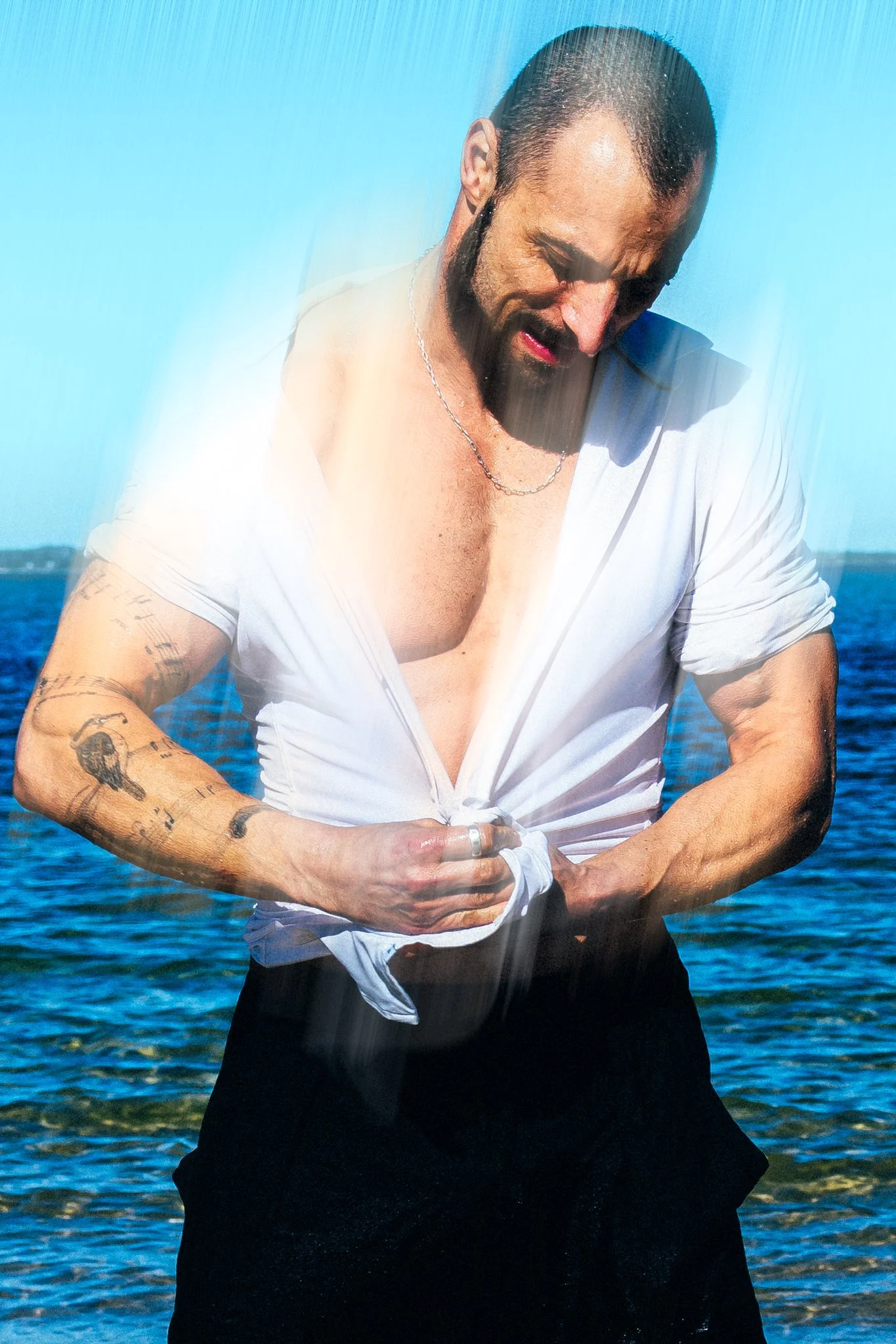 A man is standing shirtless in front of water, wearing a white shirt, which he is knotting at the waist, and soaking wet.