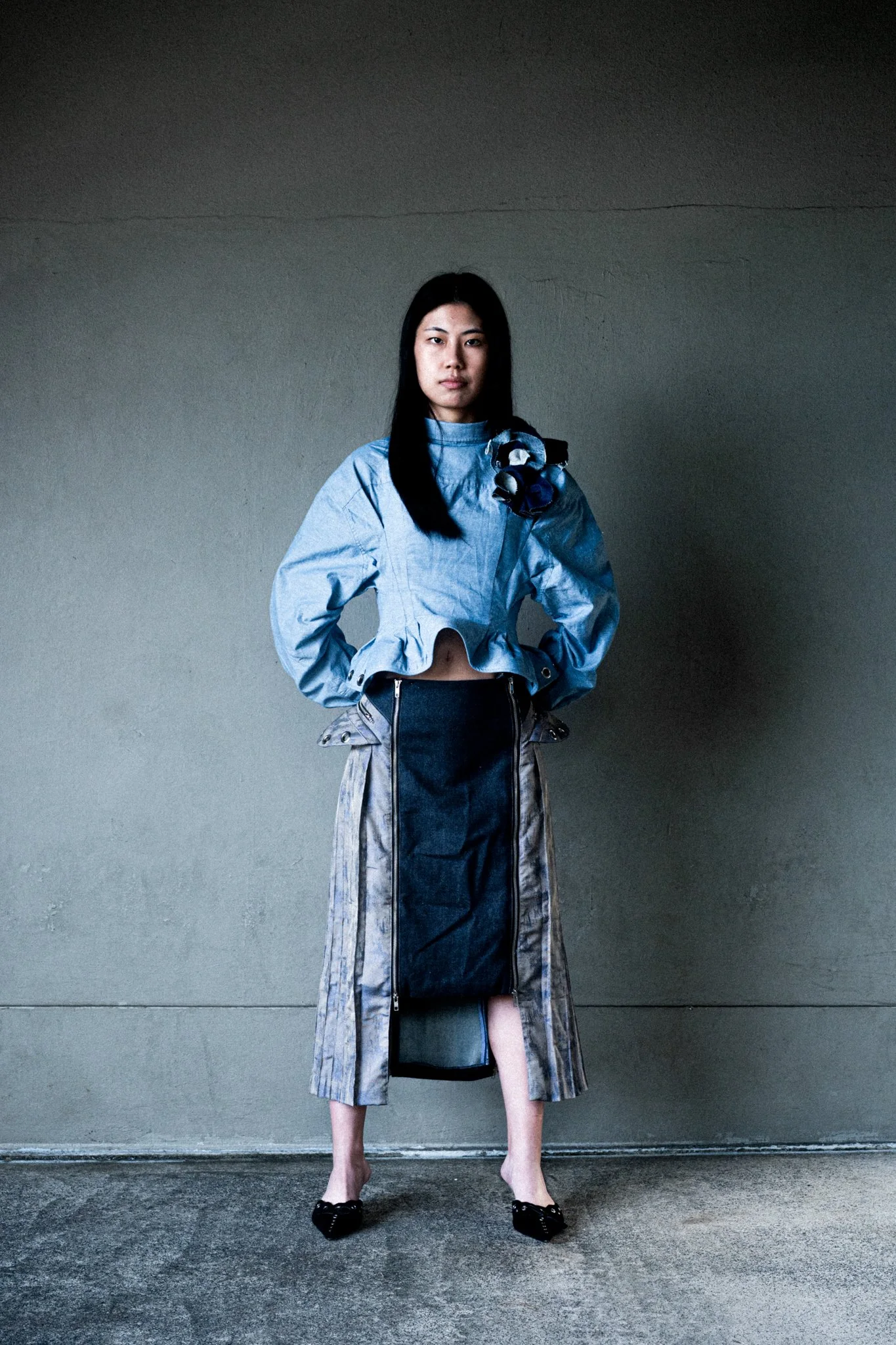 A woman with long black hair stands against a plain wall, wearing a blue cropped jacket with a decorative shoulder detail and a long skirt with zippers and a slit, paired with black pointed shoes.
