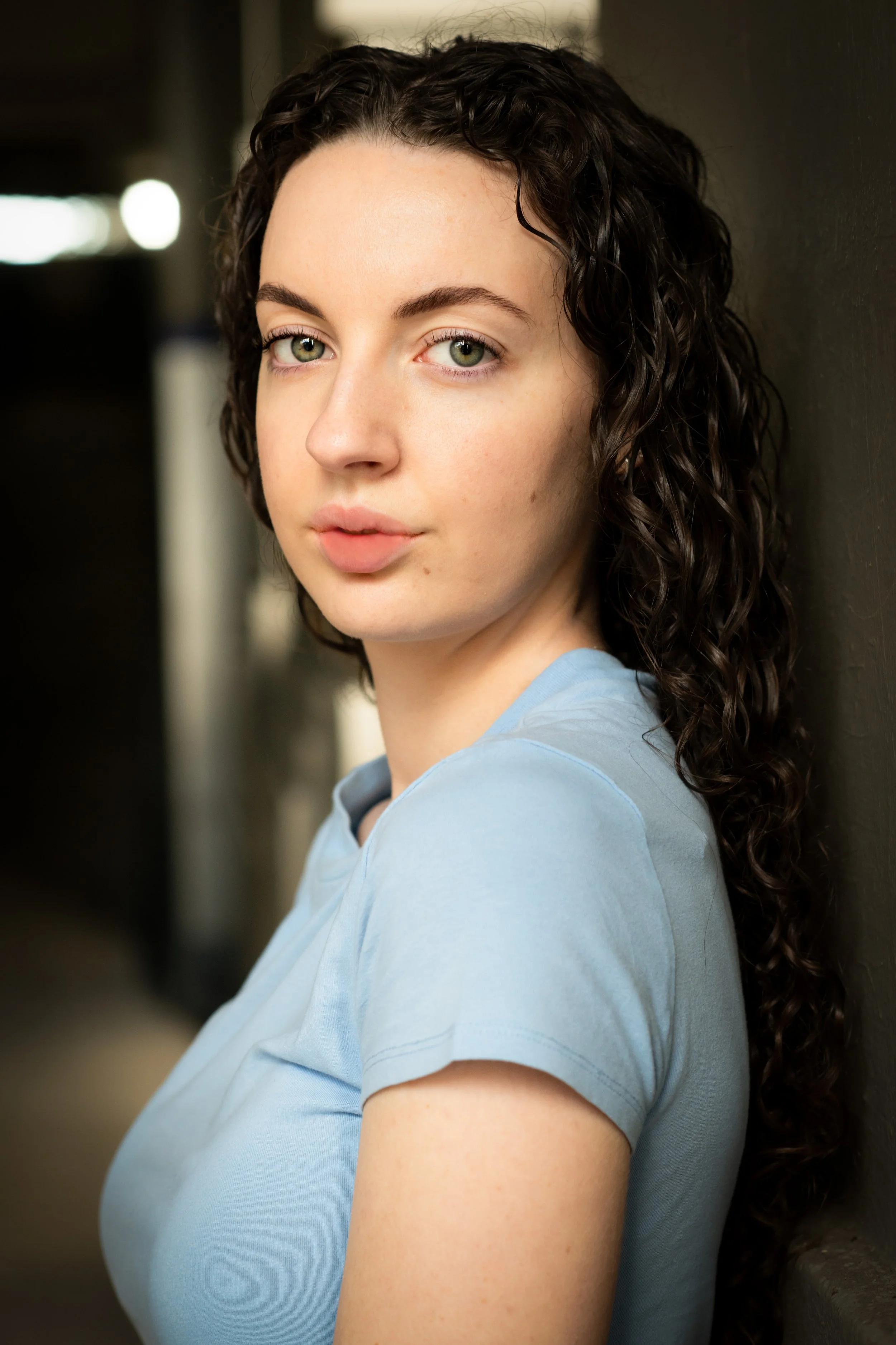 A young woman with curly dark hair and green eyes, wearing a light blue shirt, leaning against a dark wall inside a building.