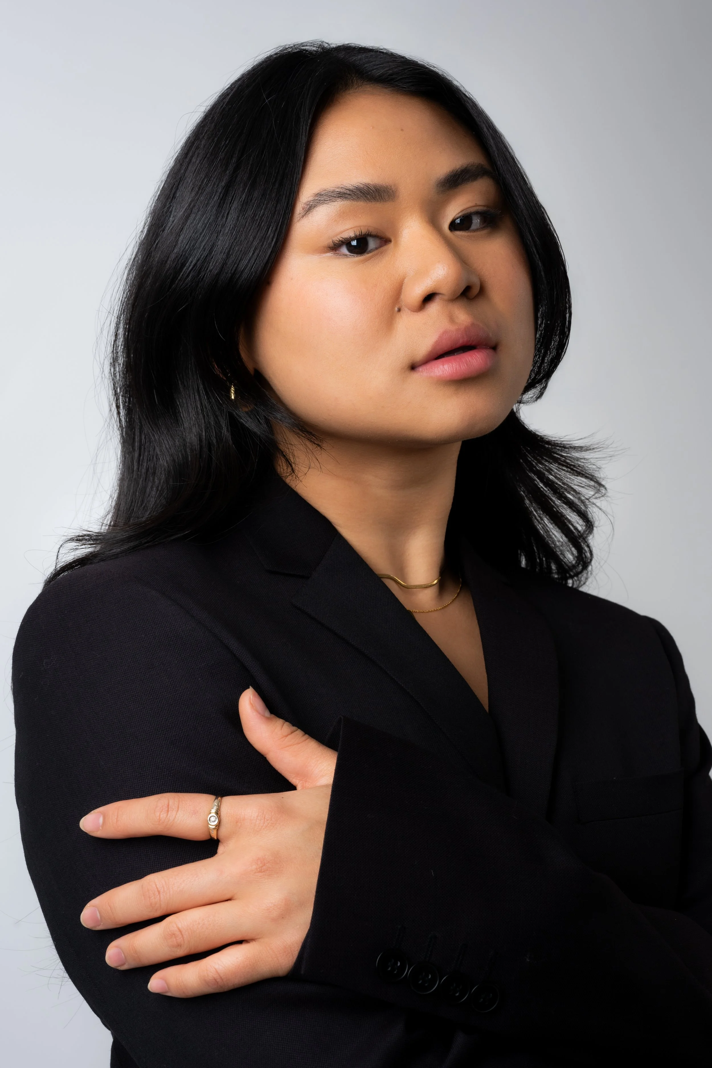A professional woman with long black hair wearing a black blazer, gold jewelry, and a neutral expression, posing with her arms crossed against a plain light gray background.