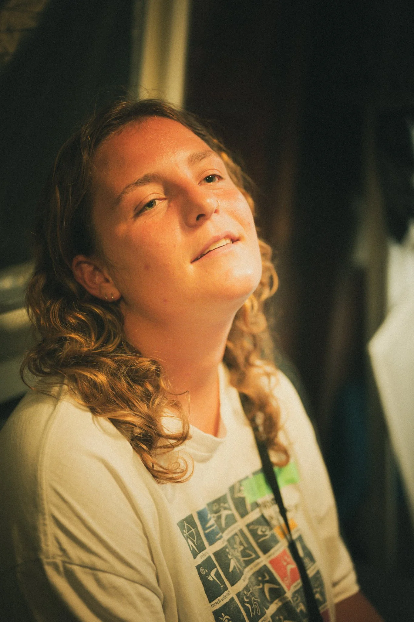 A young woman with curly light brown hair and a pierced nose, wearing a beige T-shirt with colorful graphic prints, is looking slightly upward with a relaxed expression in a dimly lit indoor setting.