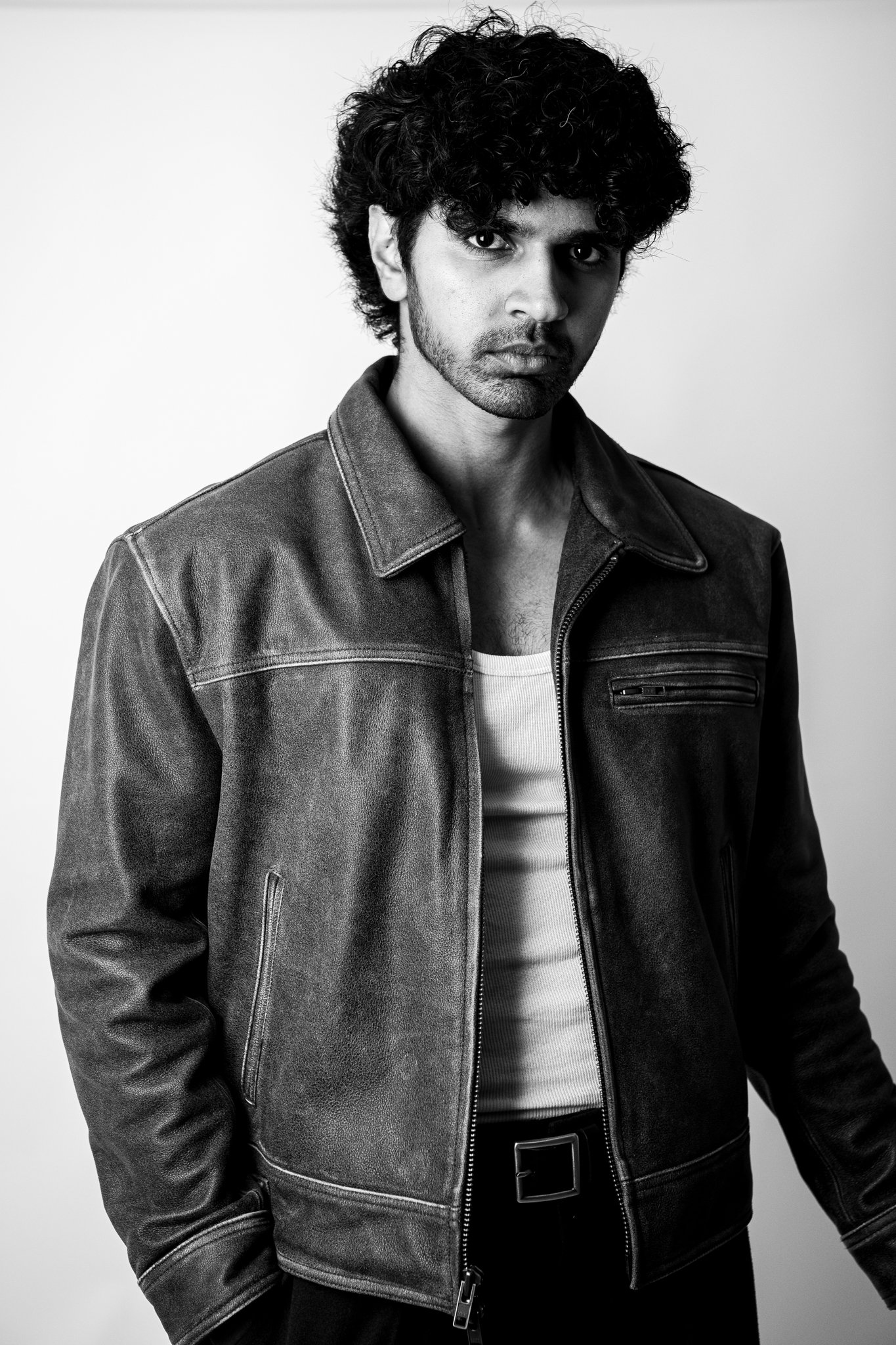 Black and white photo of a man with curly dark hair, wearing a leather jacket over a white T-shirt against a plain background.