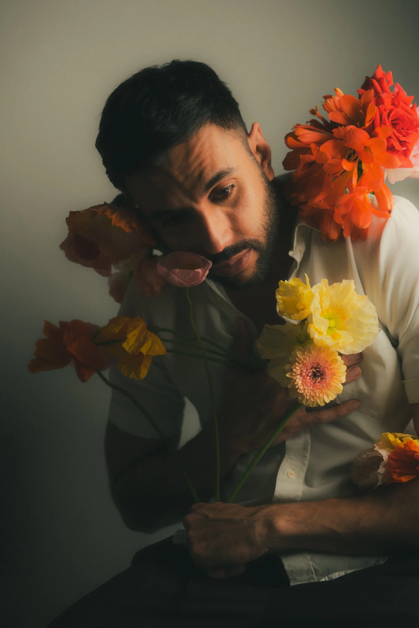A man with dark hair and a beard holding a bouquet of colorful flowers, including yellow, pink, and orange blossoms, while leaning against a plain wall.