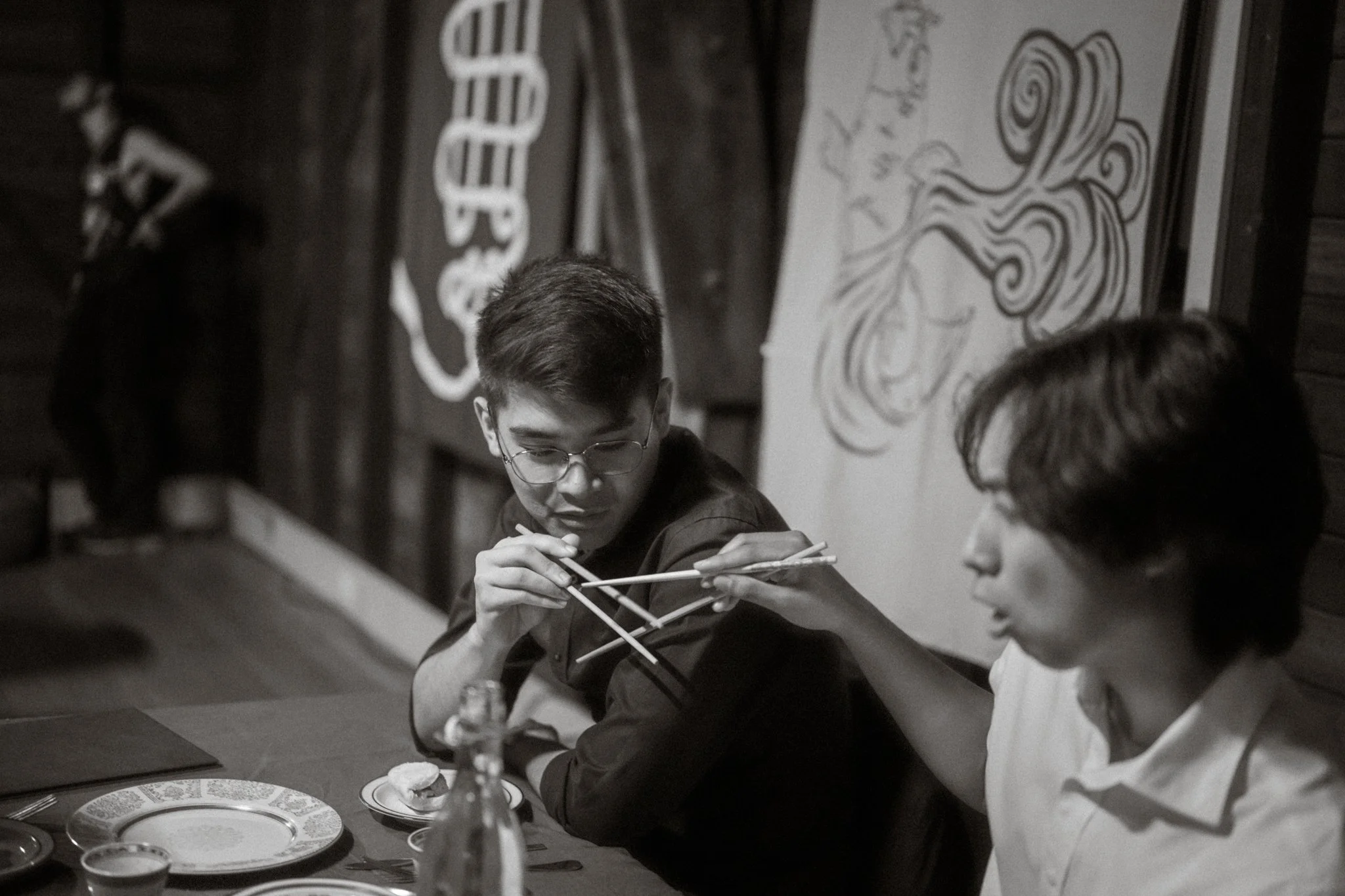 Two young men seated at a dinner table, one passing food with chopsticks to the other, in a restaurant with artistic wall decorations.