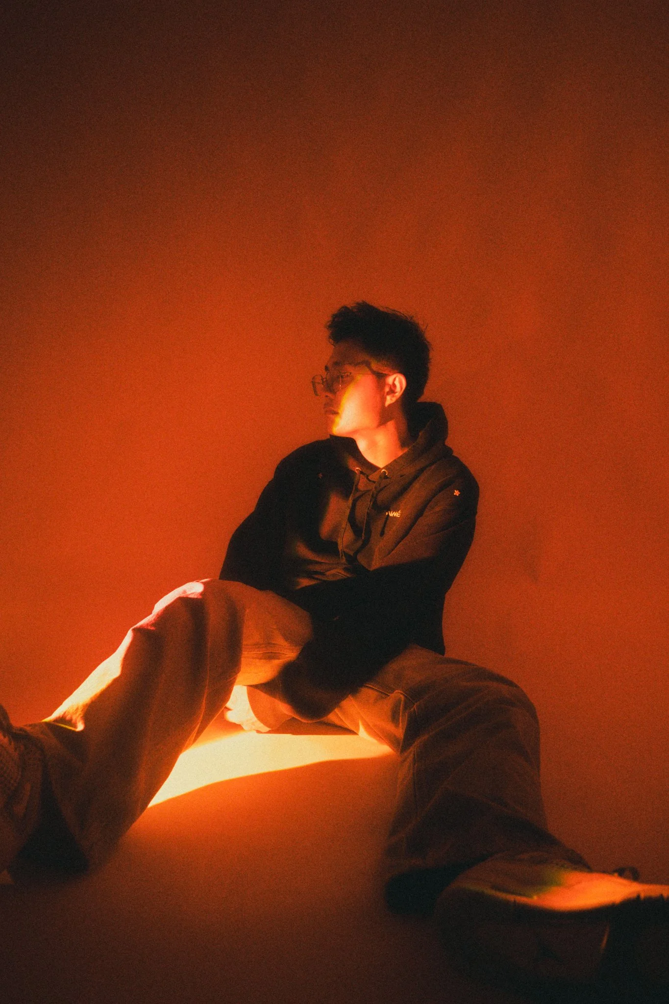 A young man sitting on the ground against an orange background, wearing glasses, a dark jacket, and beige pants, illuminated by warm lighting.