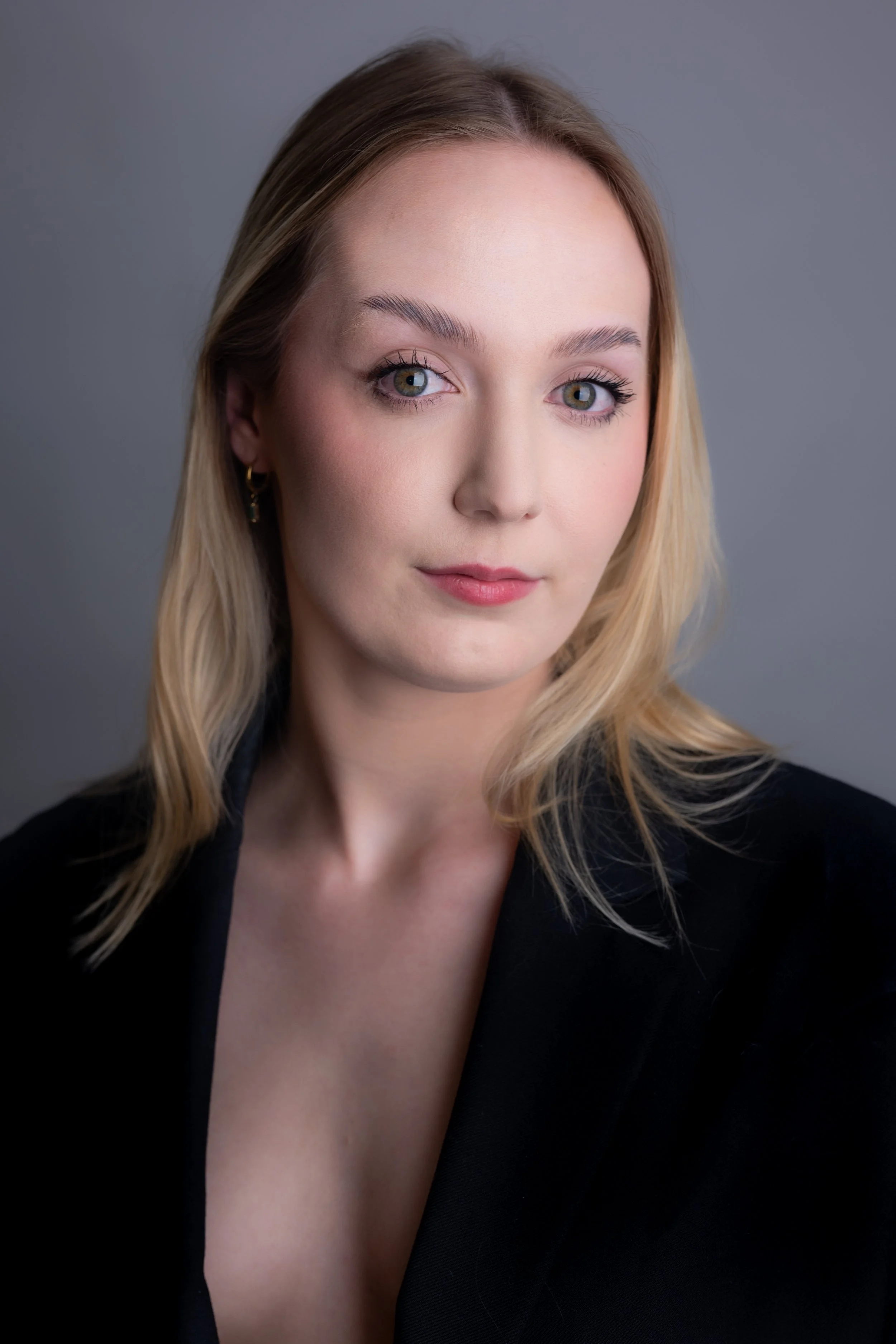 Headshot of a young woman with blonde hair and blue eyes, wearing a black blazer, against a gray background.
