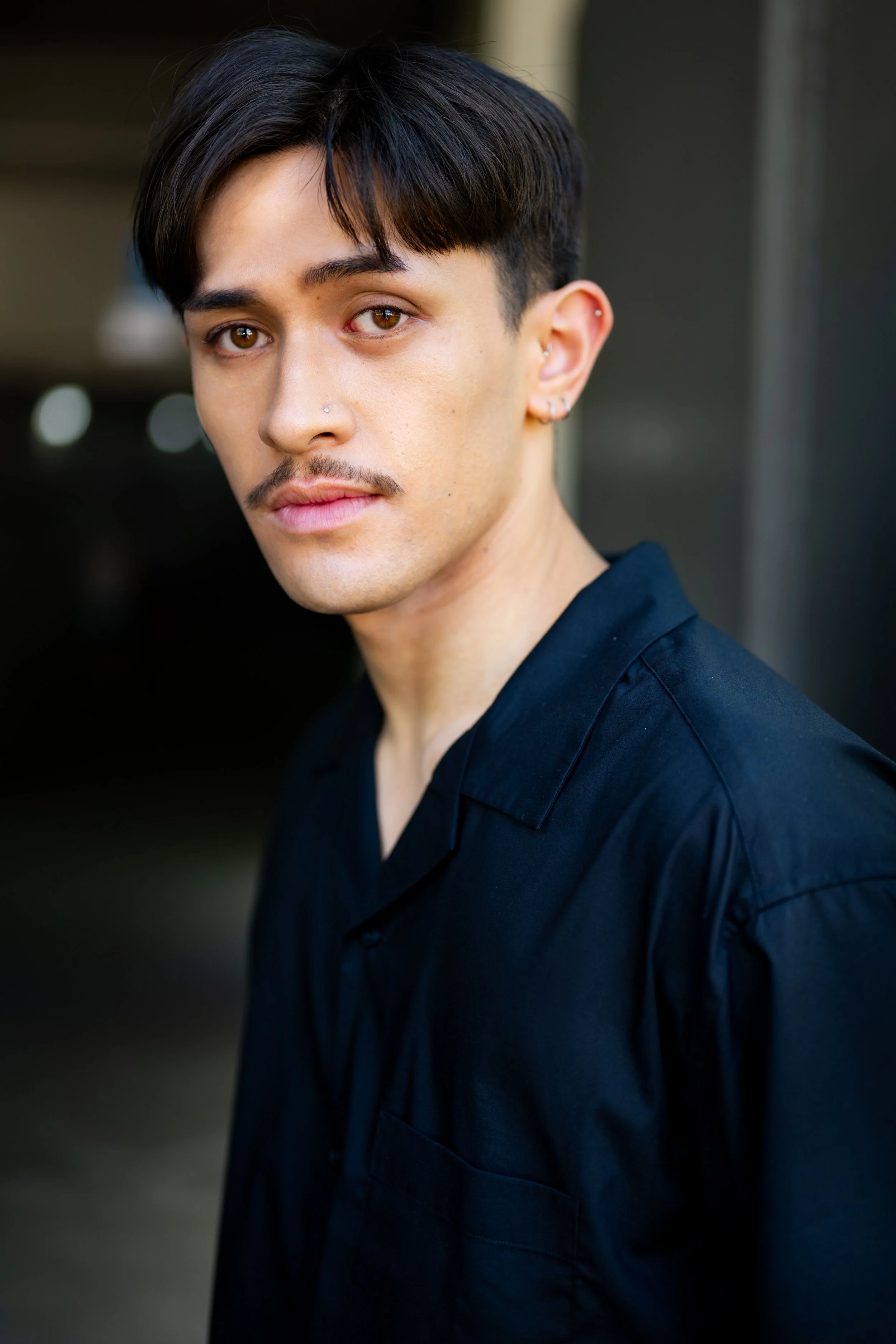 Portrait of a young man with dark hair, brown eyes, and a nose piercing, wearing a black collared shirt, outdoors.