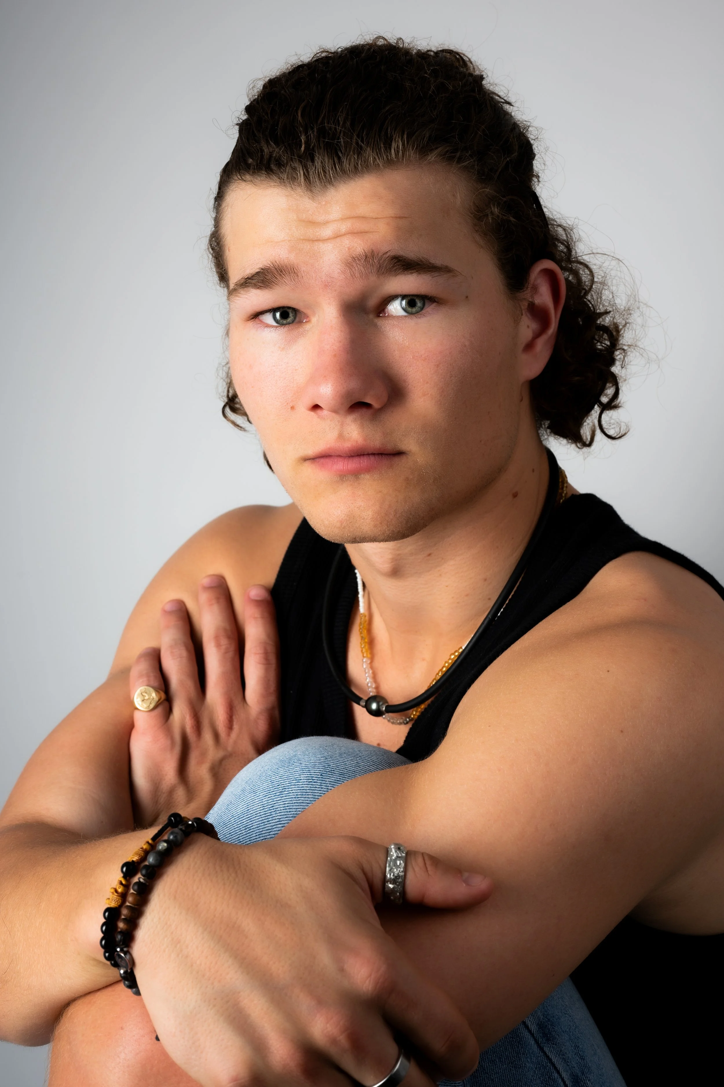 A young man with curly brown hair, blue eyes, and a serious expression is sitting with one arm crossed over his knee, resting on his shoulder. He is wearing a black sleeveless shirt, jewelry including rings, a beaded bracelet, and necklaces, against 