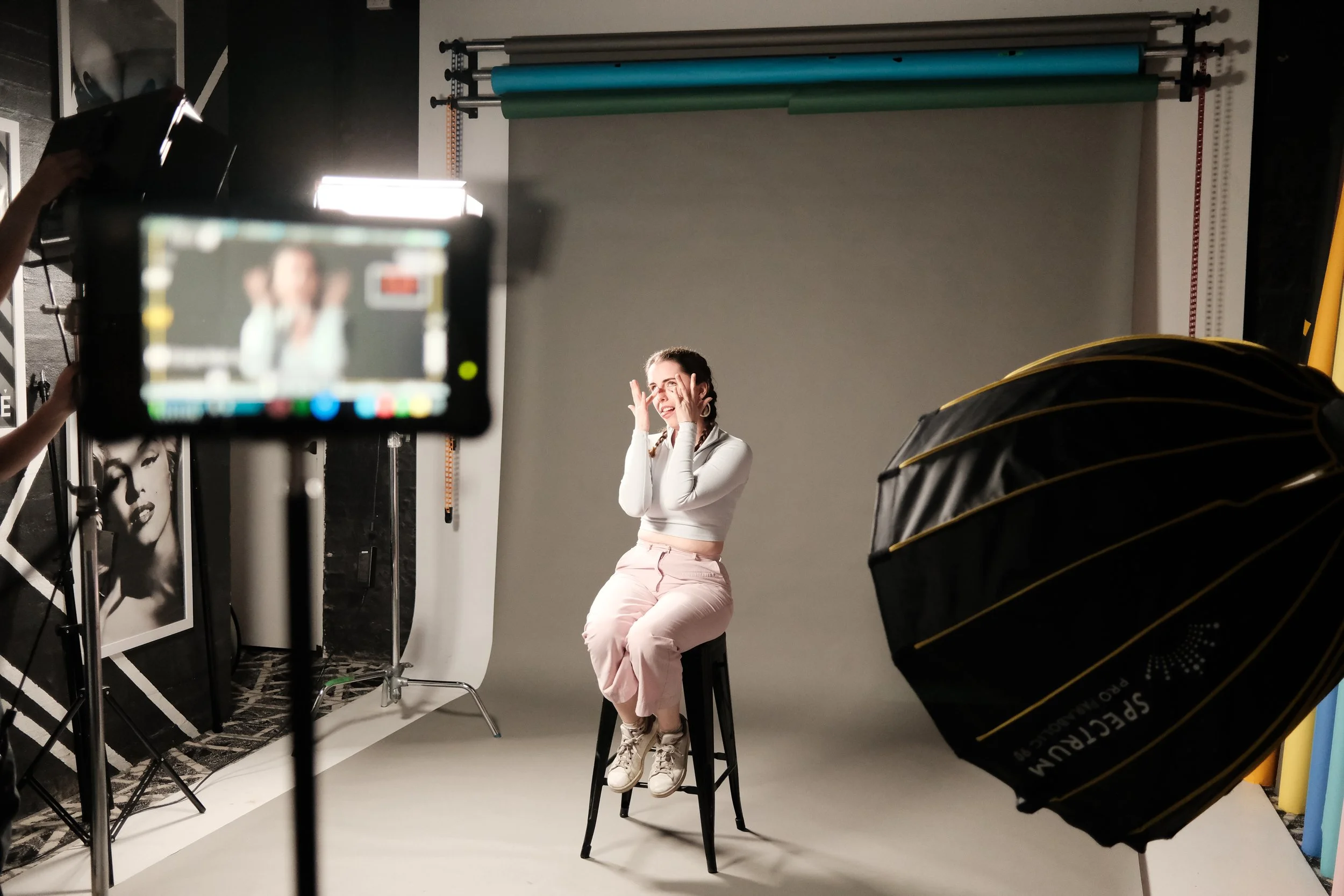 Woman sitting on a stool in a photography studio, posing with her hands near her face, while being photographed with studio equipment and a gray backdrop.