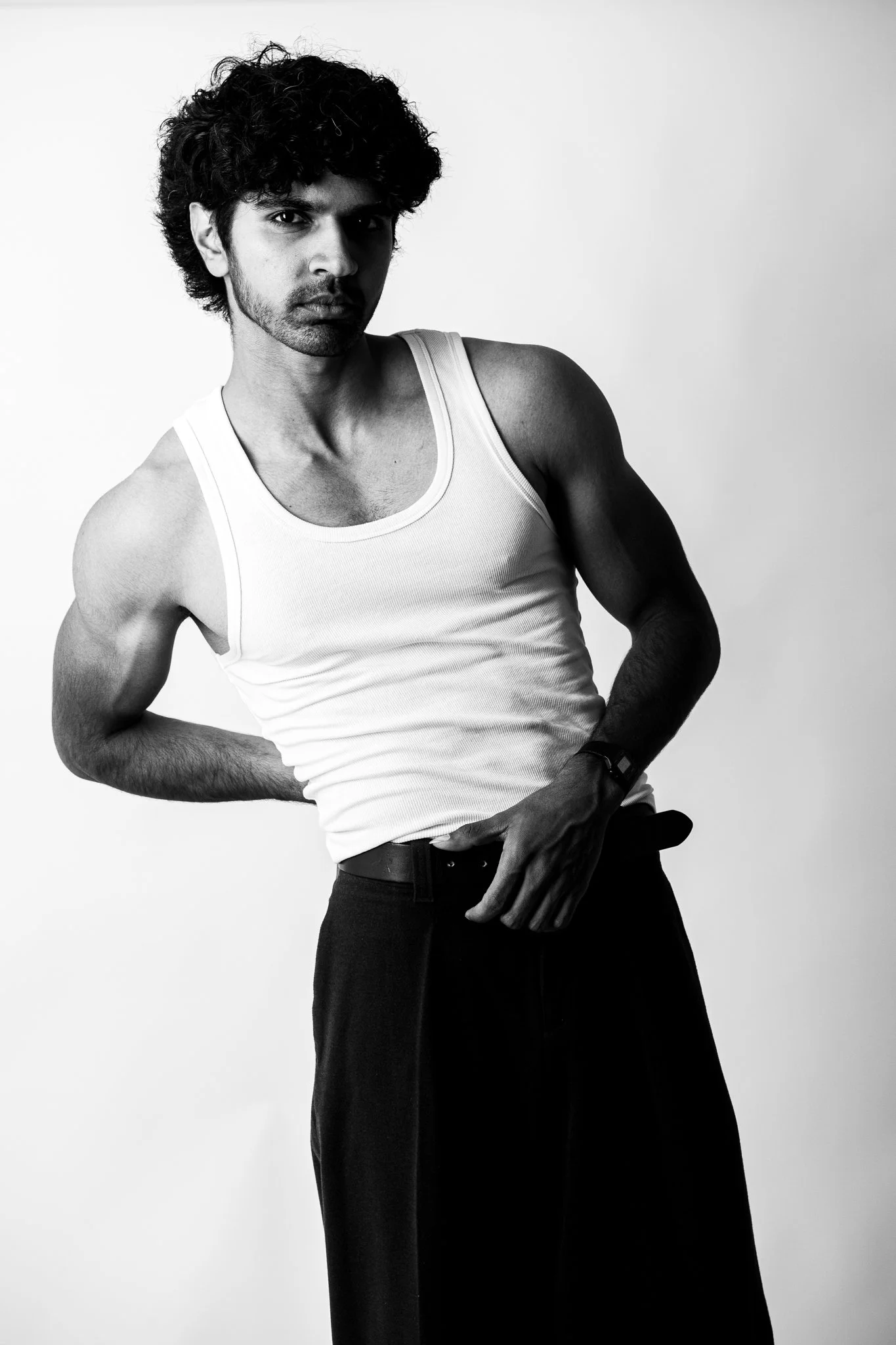 Black and white photo of a young man with curly hair wearing a white tank top and dark pants, posing against a plain background.
