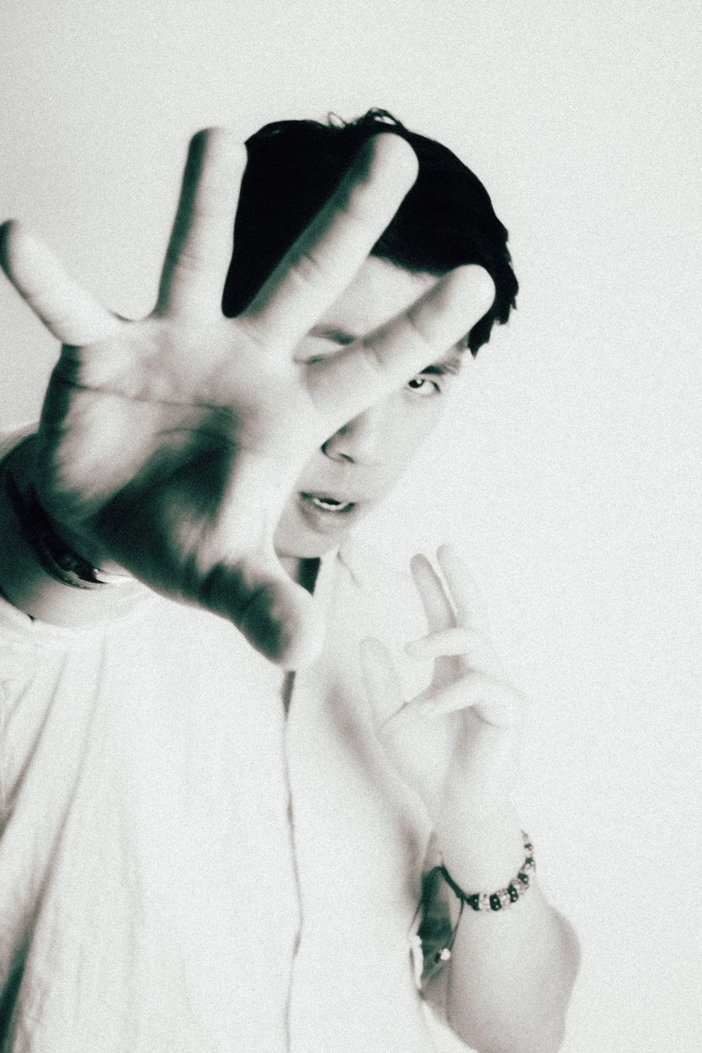 Black-and-white photo of a young man with dark hair, partially covering his face with his right hand, and making a peace sign with his left hand.