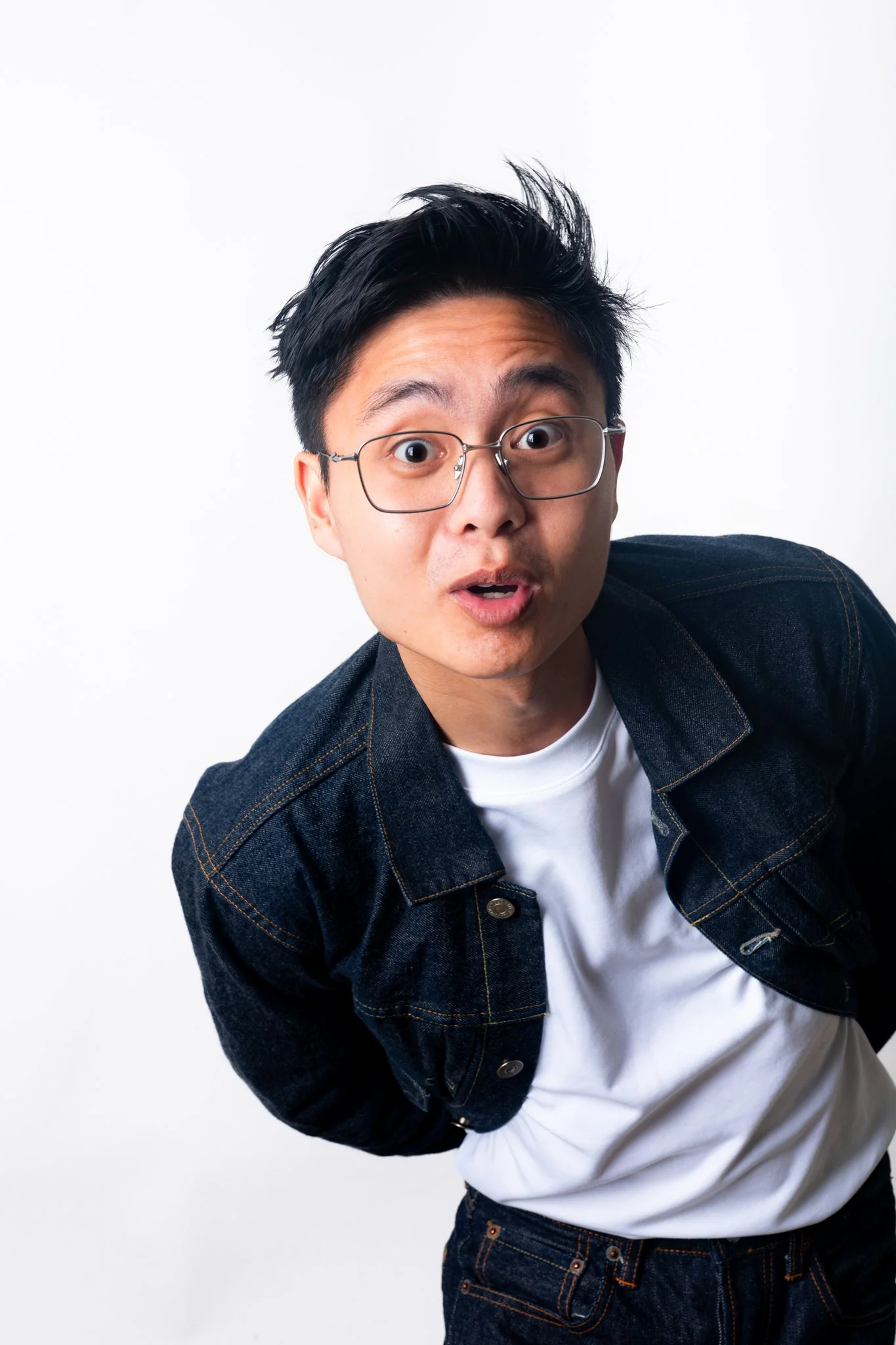 A young man with glasses and dark hair looking surprised, leaning forward against a plain white background, wearing a denim jacket over a white t-shirt.