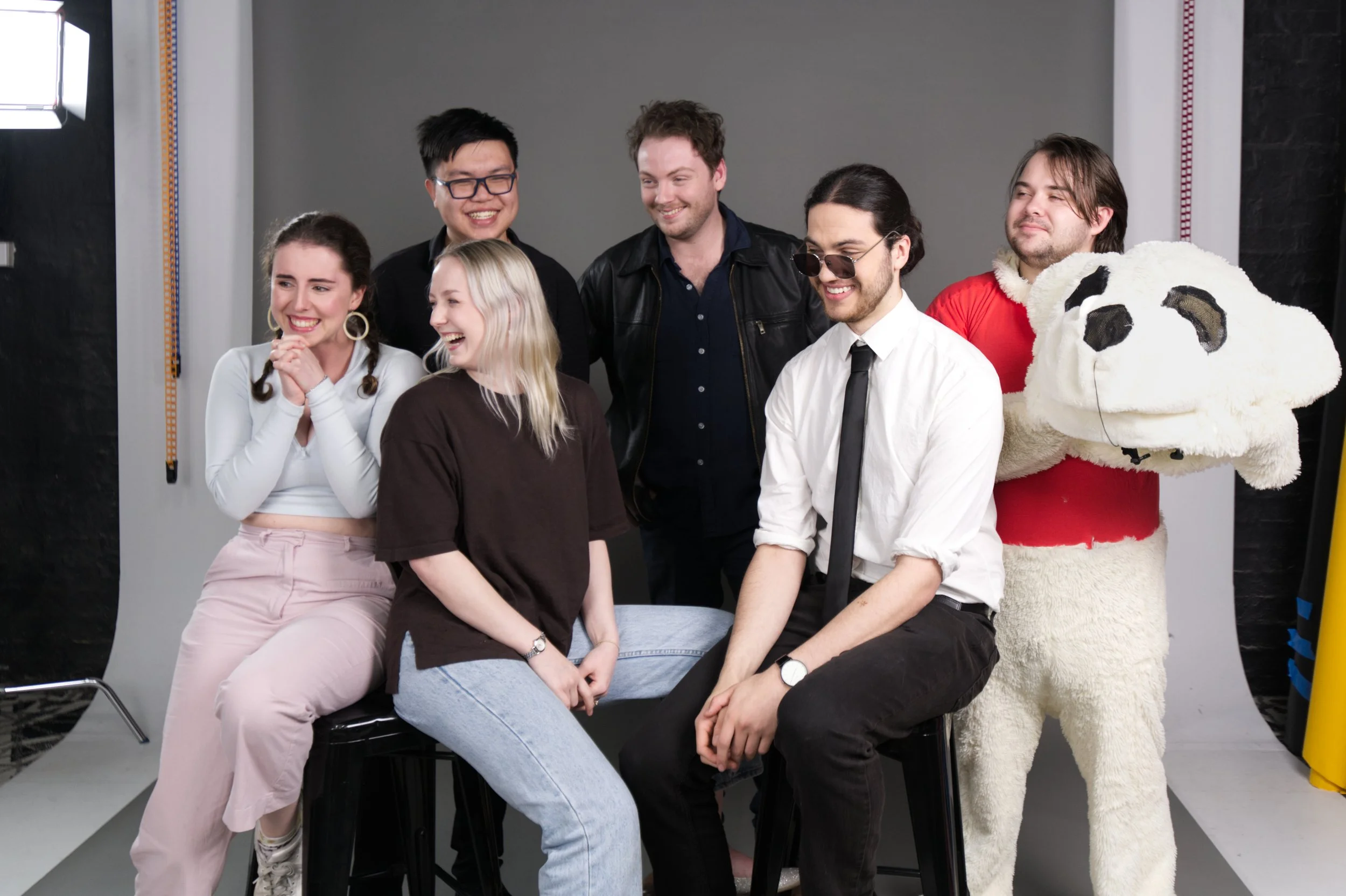 Group of seven young adults posing and laughing, with a person in a panda mascot costume holding a large panda head, in front of a grey backdrop in a photo studio.