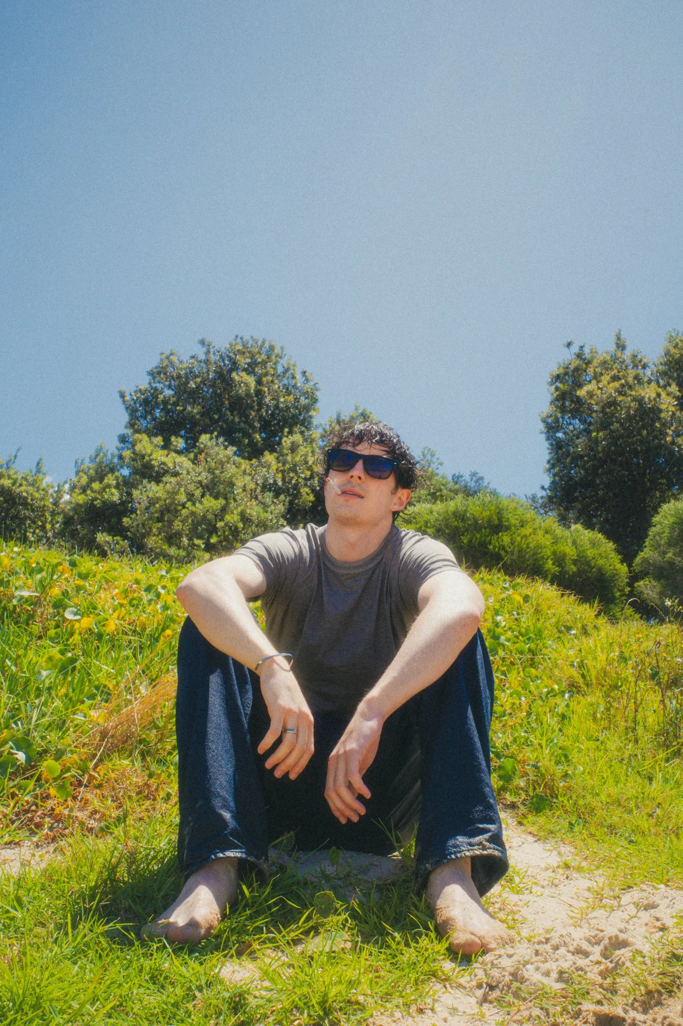 A young man with sunglasses and dark curly hair sitting barefoot on grass with soil, surrounded by green bushes and trees on a sunny day with a clear blue sky.