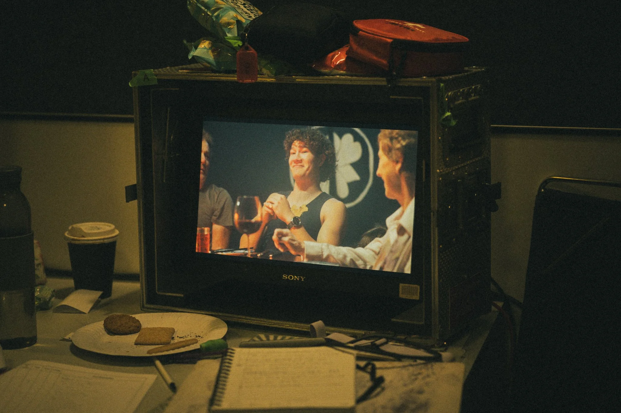 On a small table, there's a plate with cookies and a notebook with a pen. A small monitor displays a scene from a TV show or movie, with three women sitting at a table, one holding a wine glass, involved in a conversation. The surroundings are dimly 