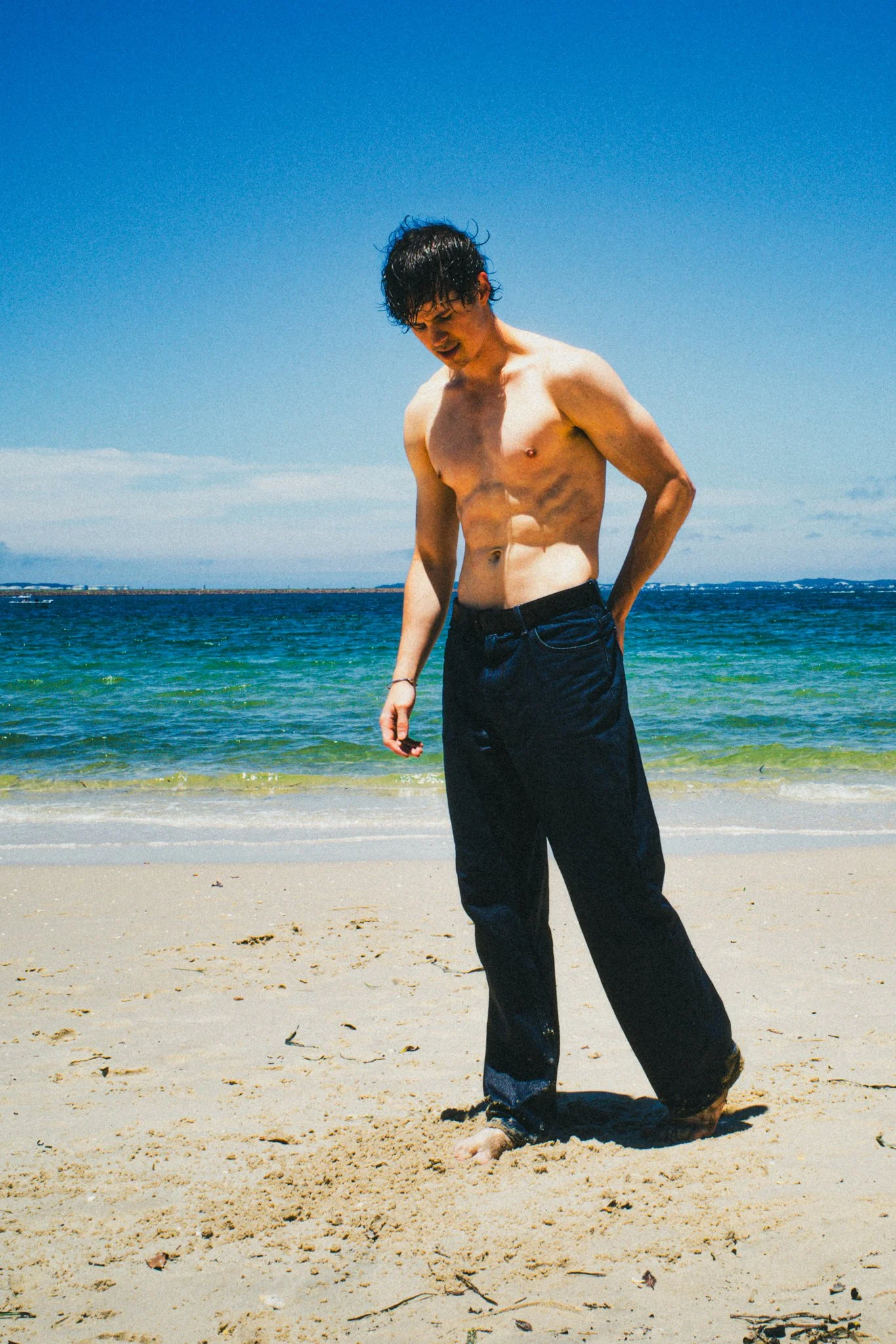 A shirtless young man with dark, wet hair standing barefoot on a sandy beach with waves and blue sky in the background.