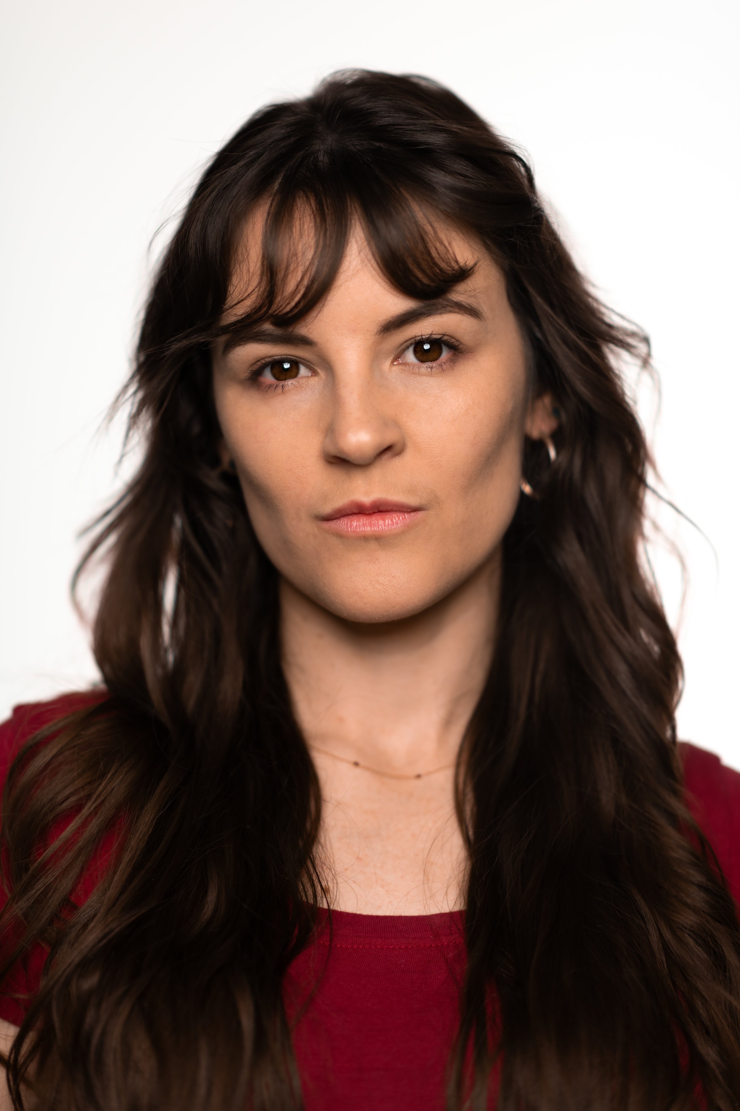 A woman with brown hair and brown eyes looking directly at the camera, wearing a red top and a small necklace, with earrings and a neutral expression.