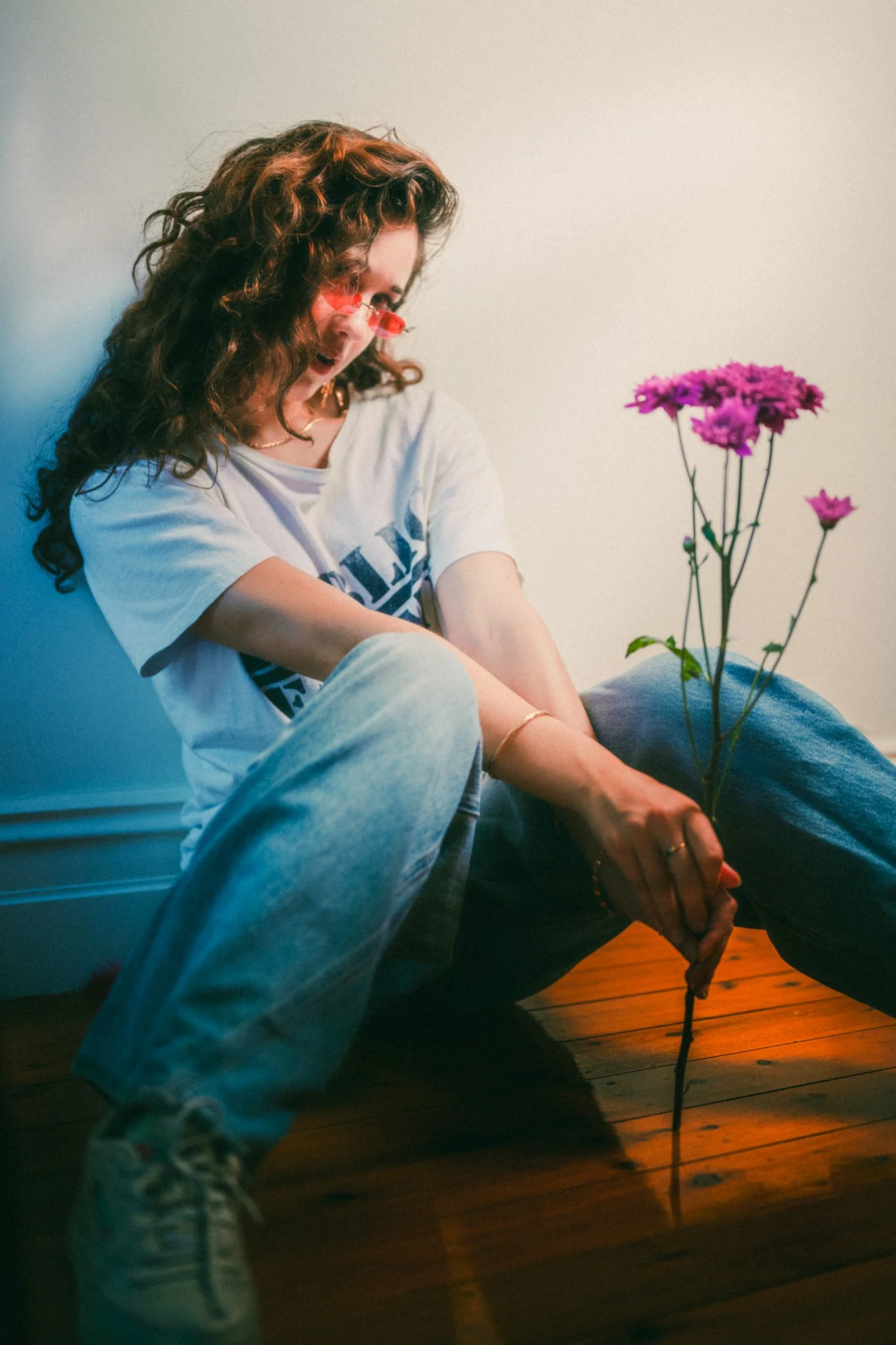 A young woman with curly red hair, wearing glasses, a white T-shirt, and jeans, sitting on a wooden floor, holding pink flowers, and smiling.