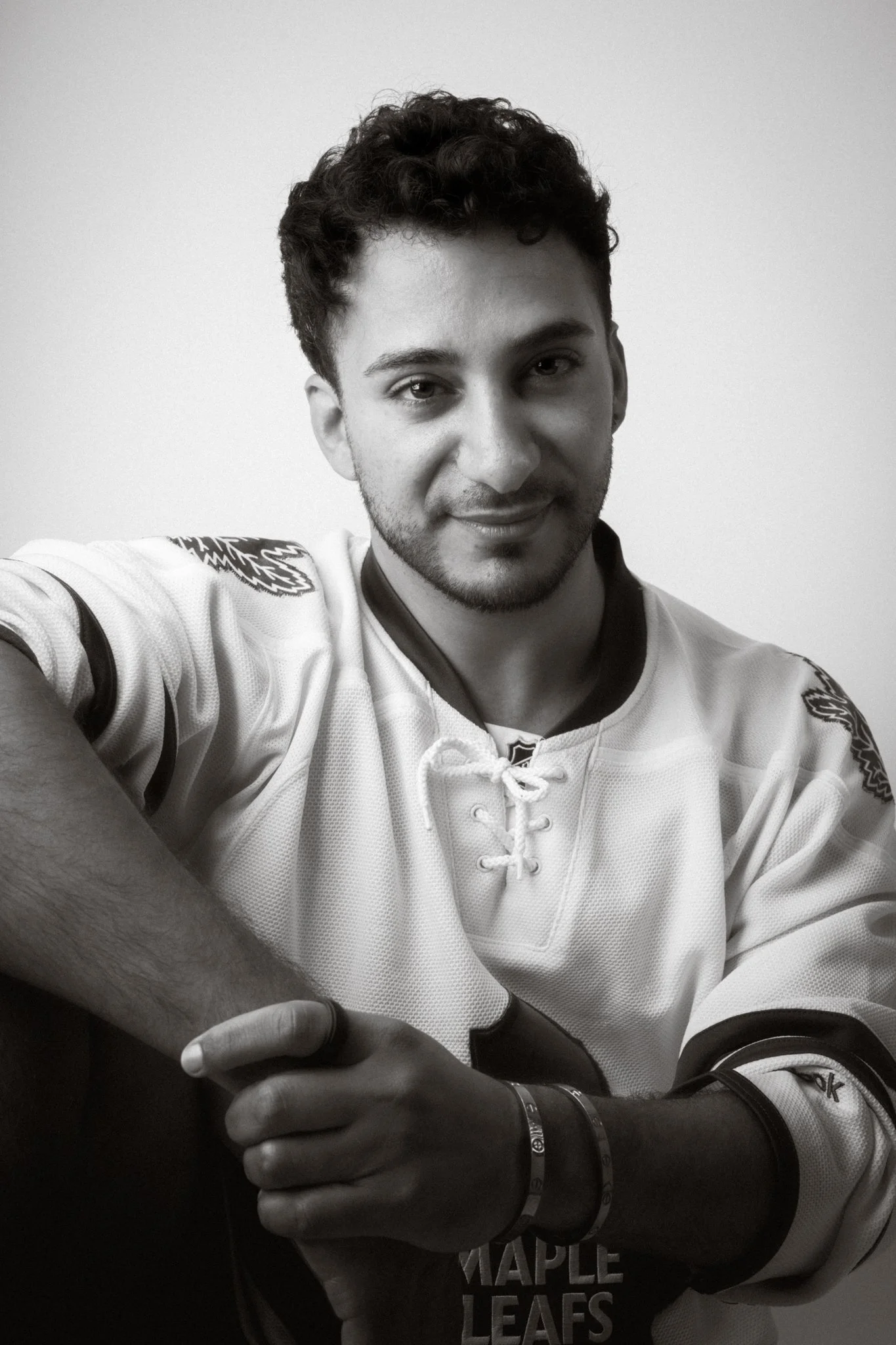 A young man with curly hair, smiling, wearing a sports jersey with Toronto Maple Leafs logo, taking a selfie against a plain background.