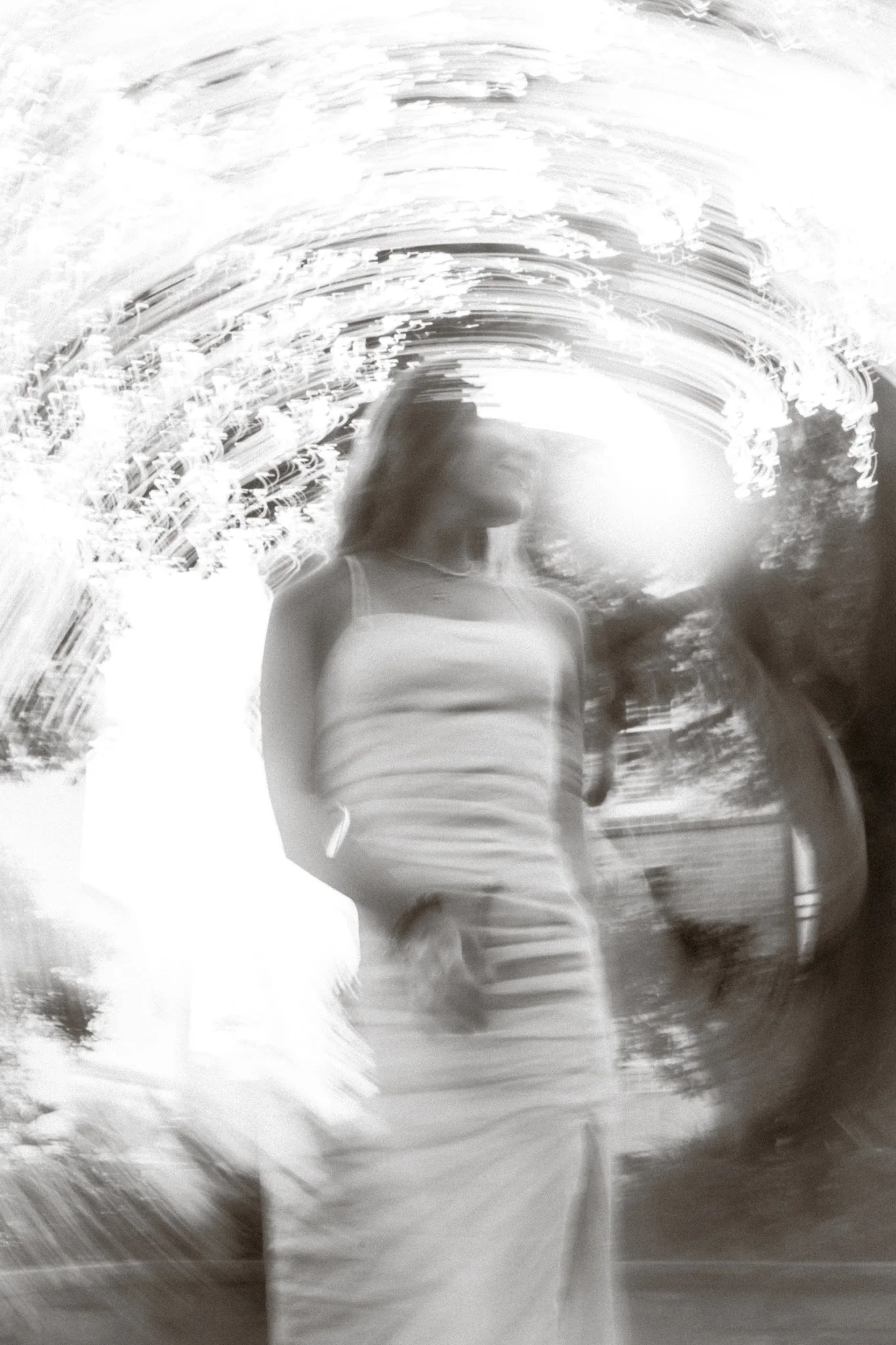 A woman in a sleeveless top and striped skirt stands outdoors at sunset or sunrise, with a blurry, abstract background and light streaks creating a swirling effect.