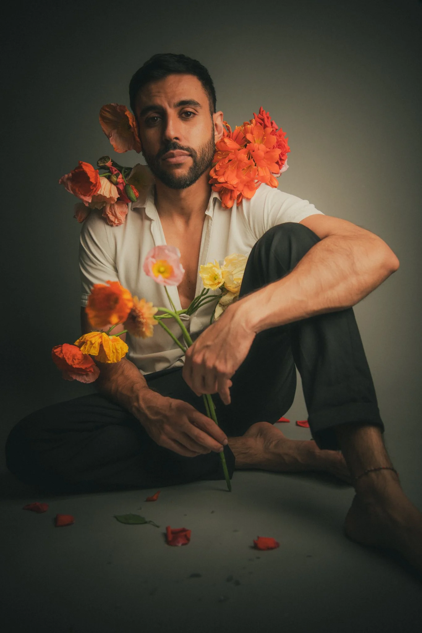 A man sitting cross-legged on the floor with flowers around him and on his shoulders, holding a flower in his hand, against a plain background.