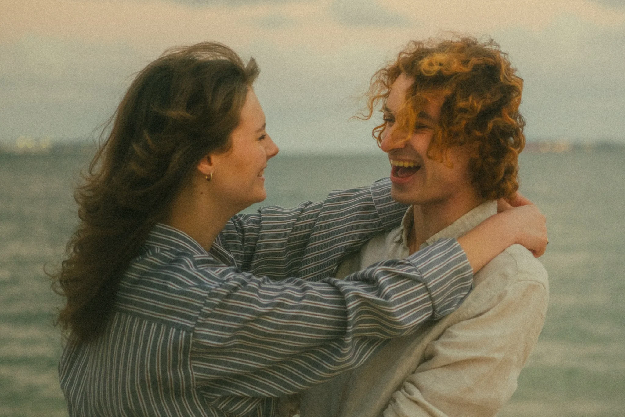 A smiling couple hugging at the beach during sunset, with the ocean in the background.