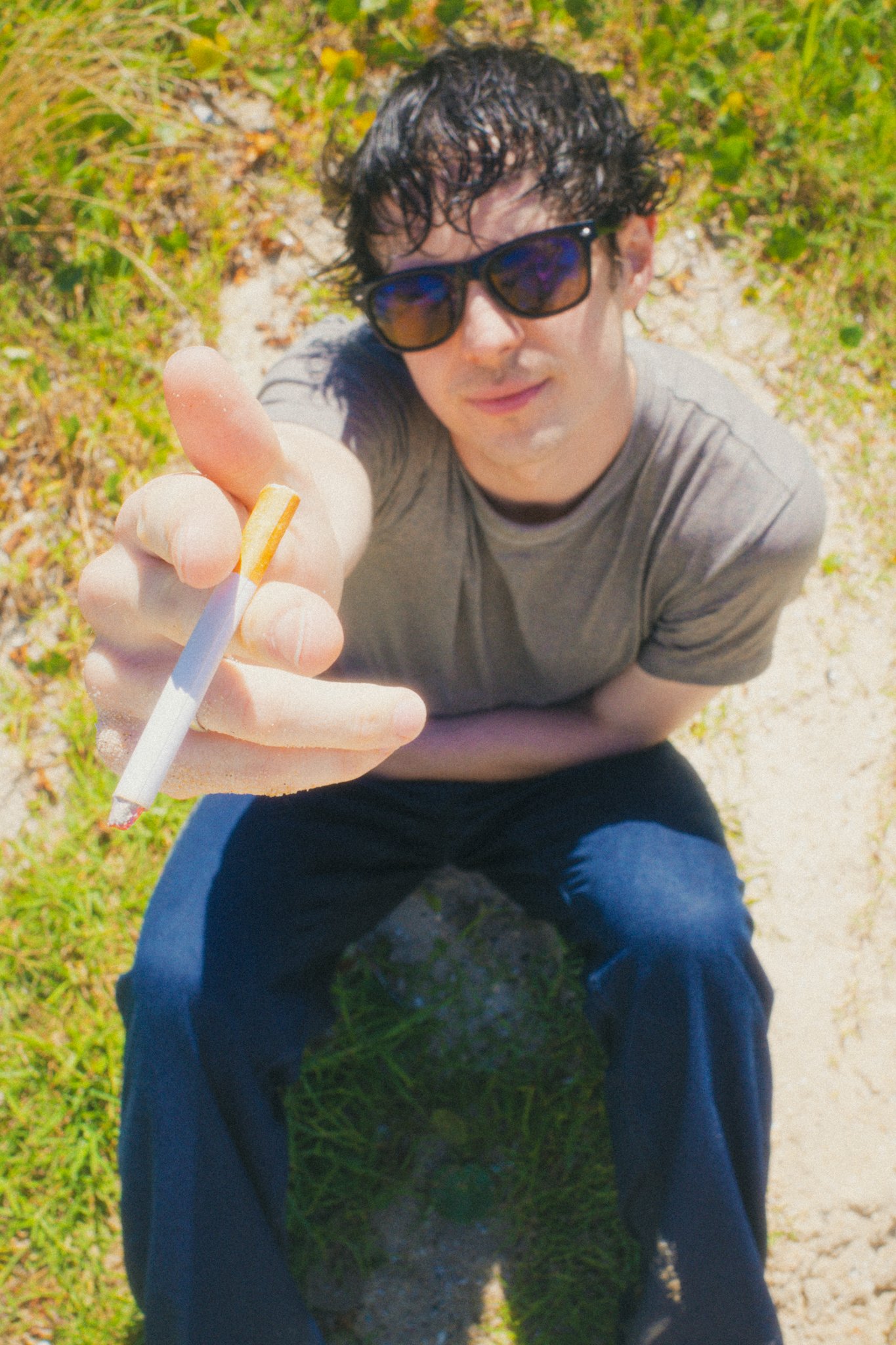 A young man with dark, curly hair and wearing sunglasses, gray t-shirt, and blue jeans, holding a cigarette extended towards the camera while sitting on a grassy and sandy outdoor path.
