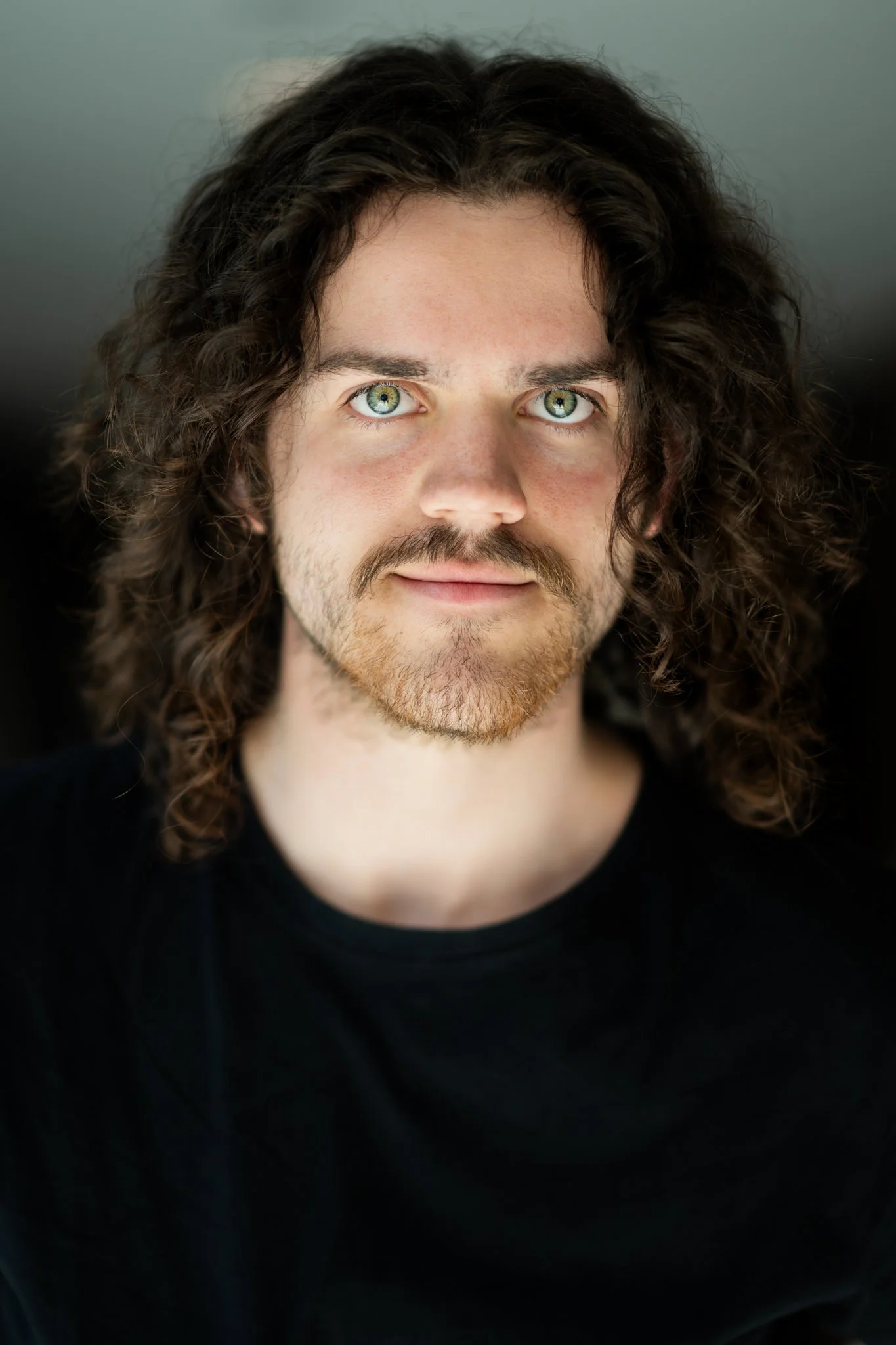 A young man with curly brown hair, green eyes, and a light beard, wearing a black shirt, looking directly into the camera.