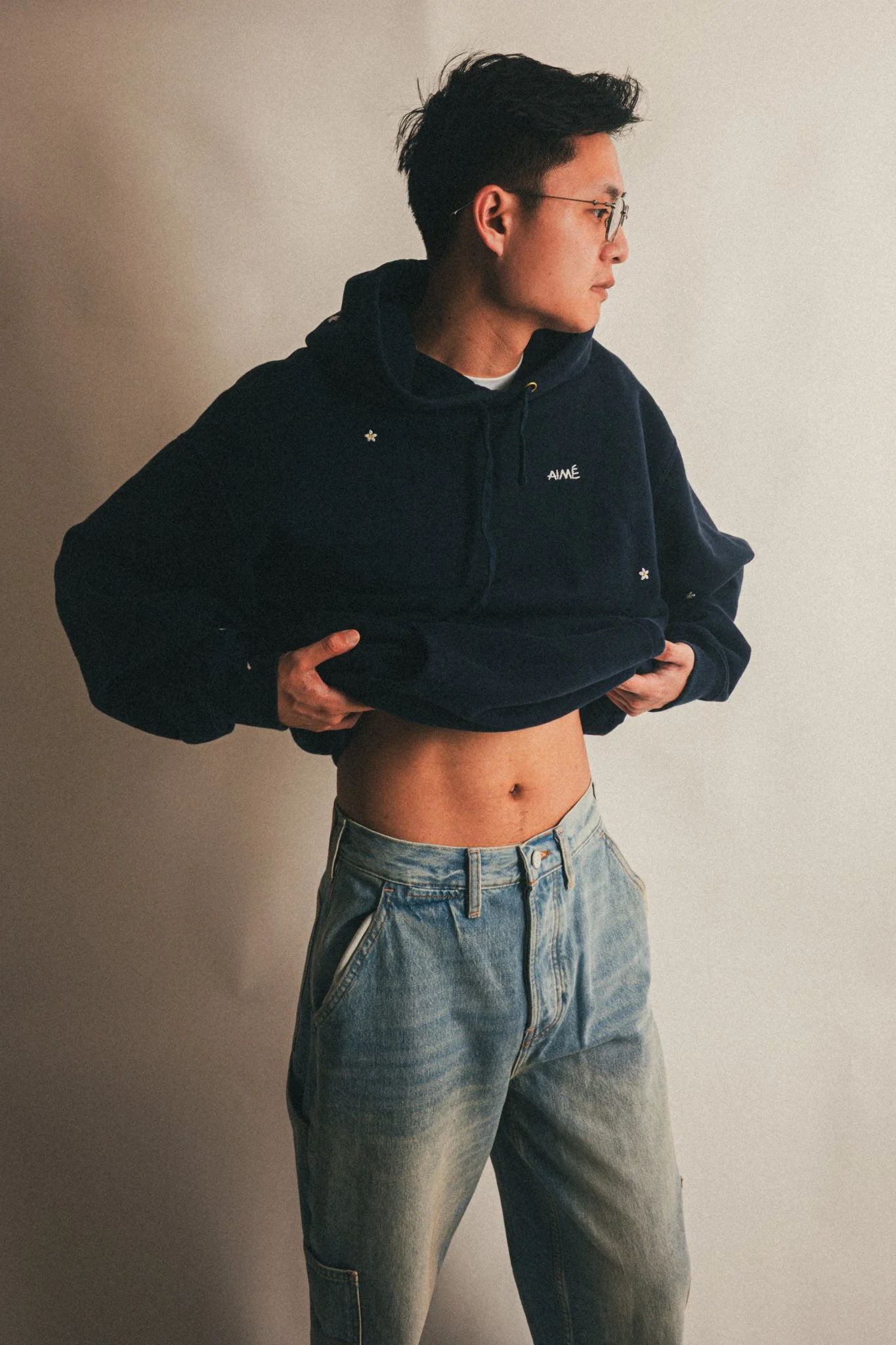 Person lifting a navy hoodie to reveal their midriff, wearing glasses and light blue jeans, standing against a plain wall.