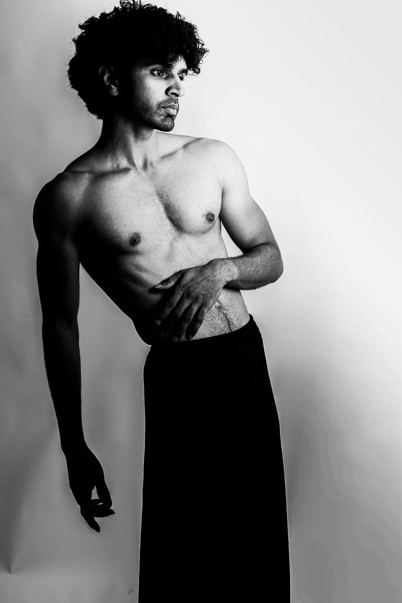 Black and white photo of a shirtless man with curly hair, looking to his left. He is standing, with his right arm hanging down and his left arm resting on his stomach, with his hand covering part of his abdomen. The background is plain and light-colo