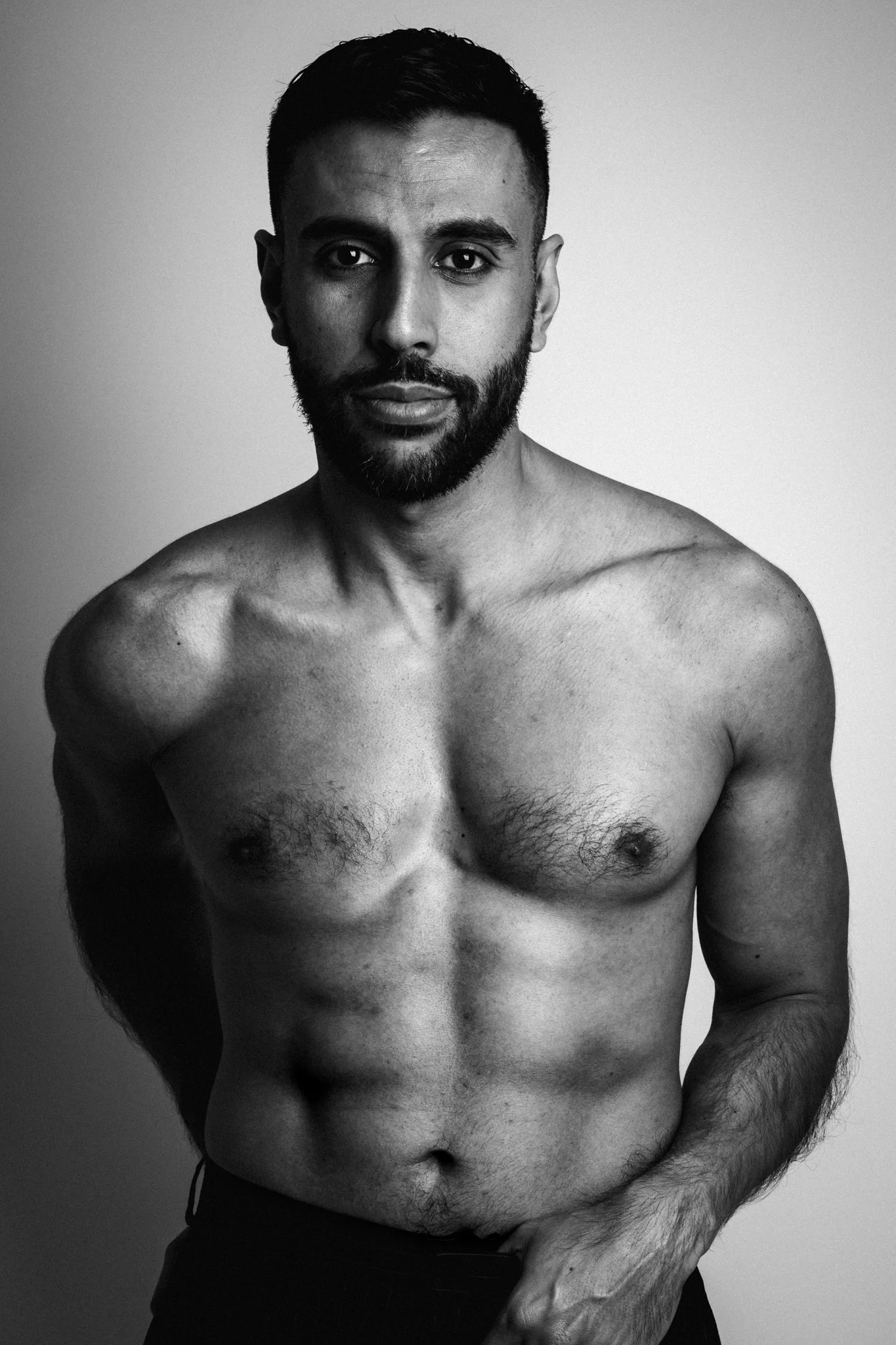Black and white portrait of a shirtless man with a beard, looking directly at the camera against a plain background.