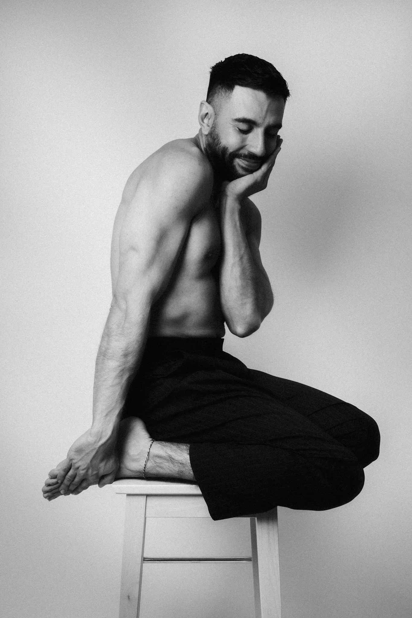 A black and white photo of a shirtless man sitting cross-legged on a stool, with his eyes closed and one hand resting on his cheek, appearing to be in a state of contemplation or serenity.