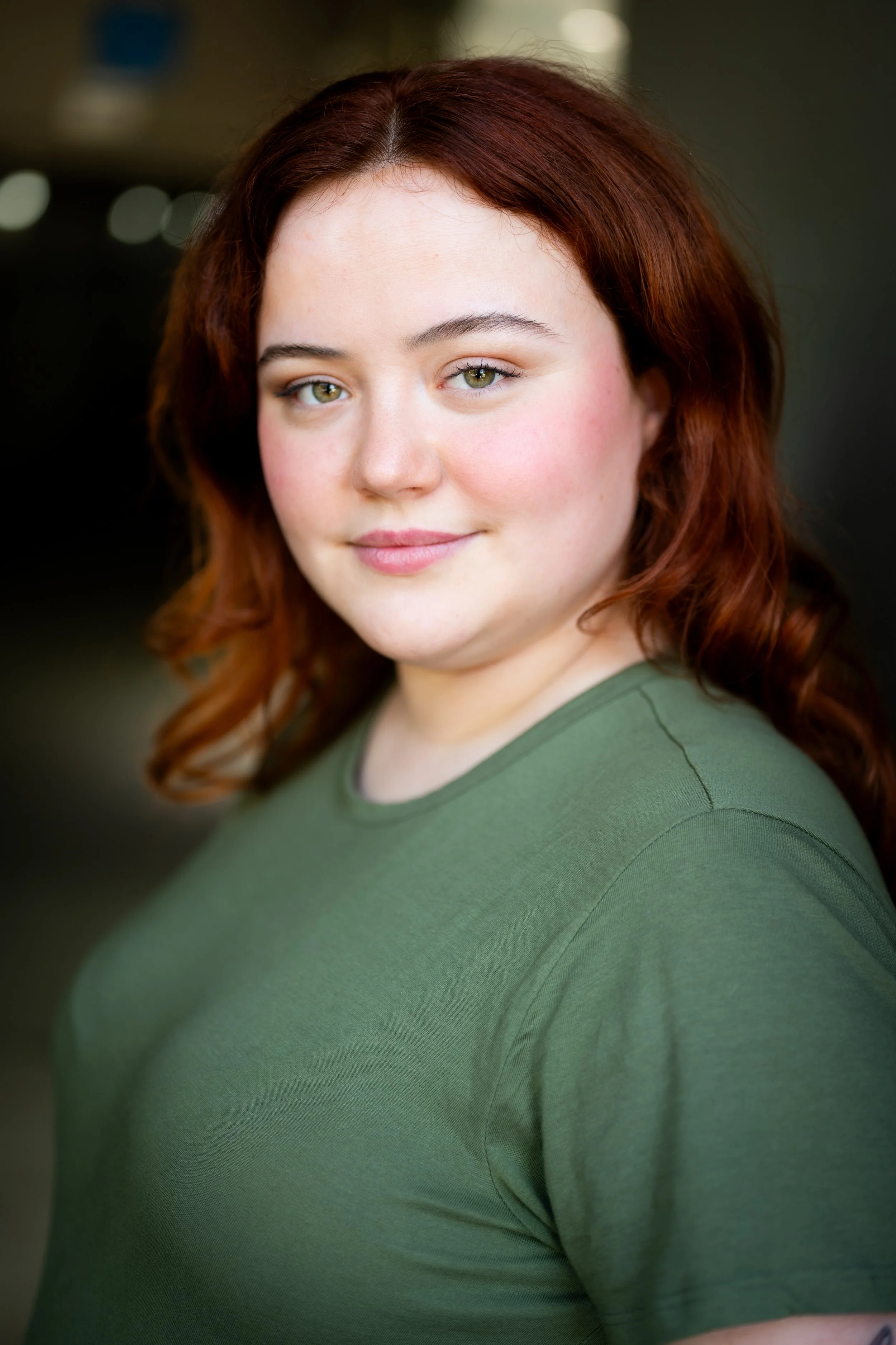 A young woman with red hair, fair skin, and green eyes wearing a green shirt. She is looking at the camera with a slight smile. The background is blurred.