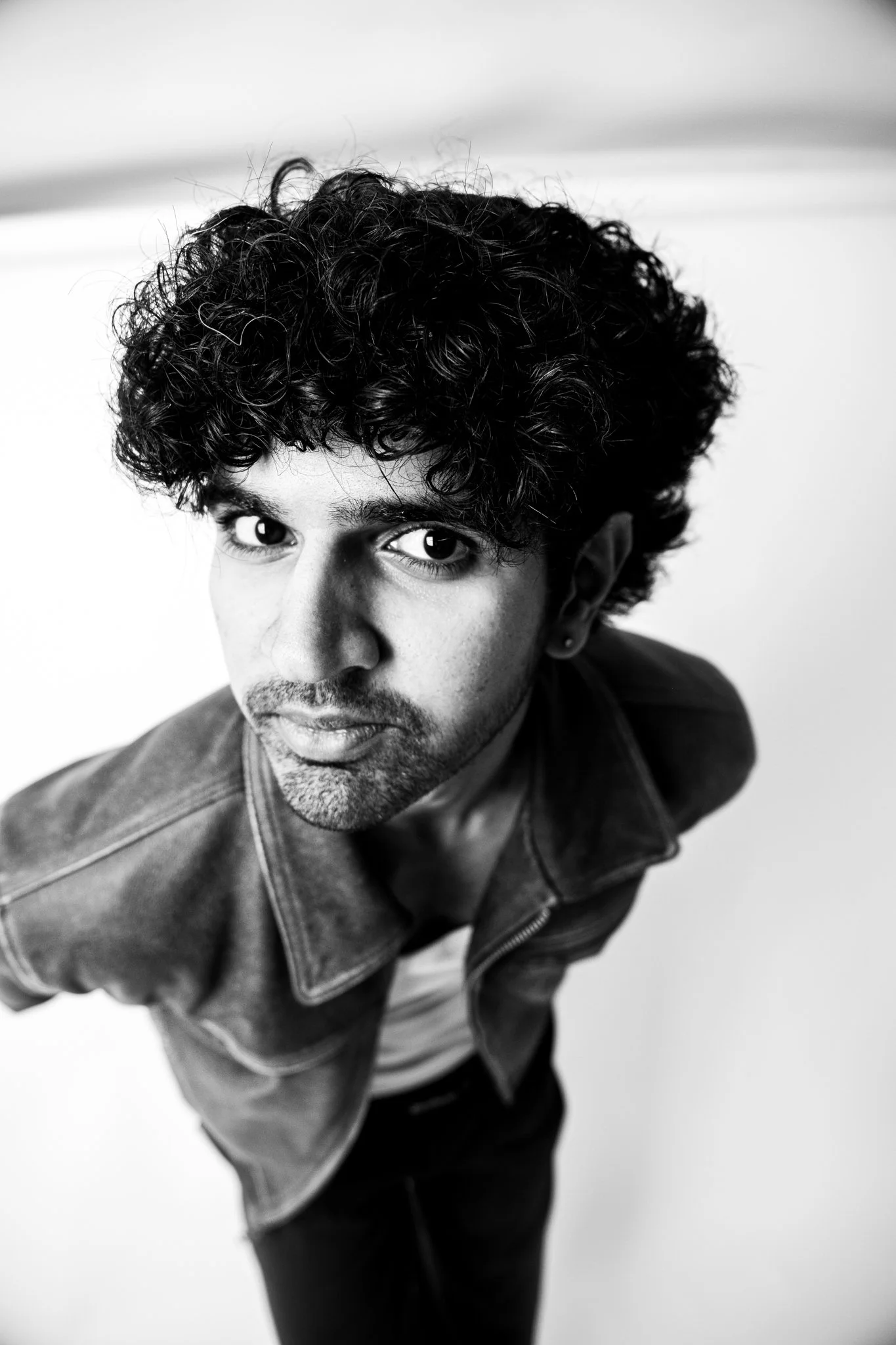 Black and white portrait of a man with curly hair, wearing a denim jacket, looking upward at the camera.