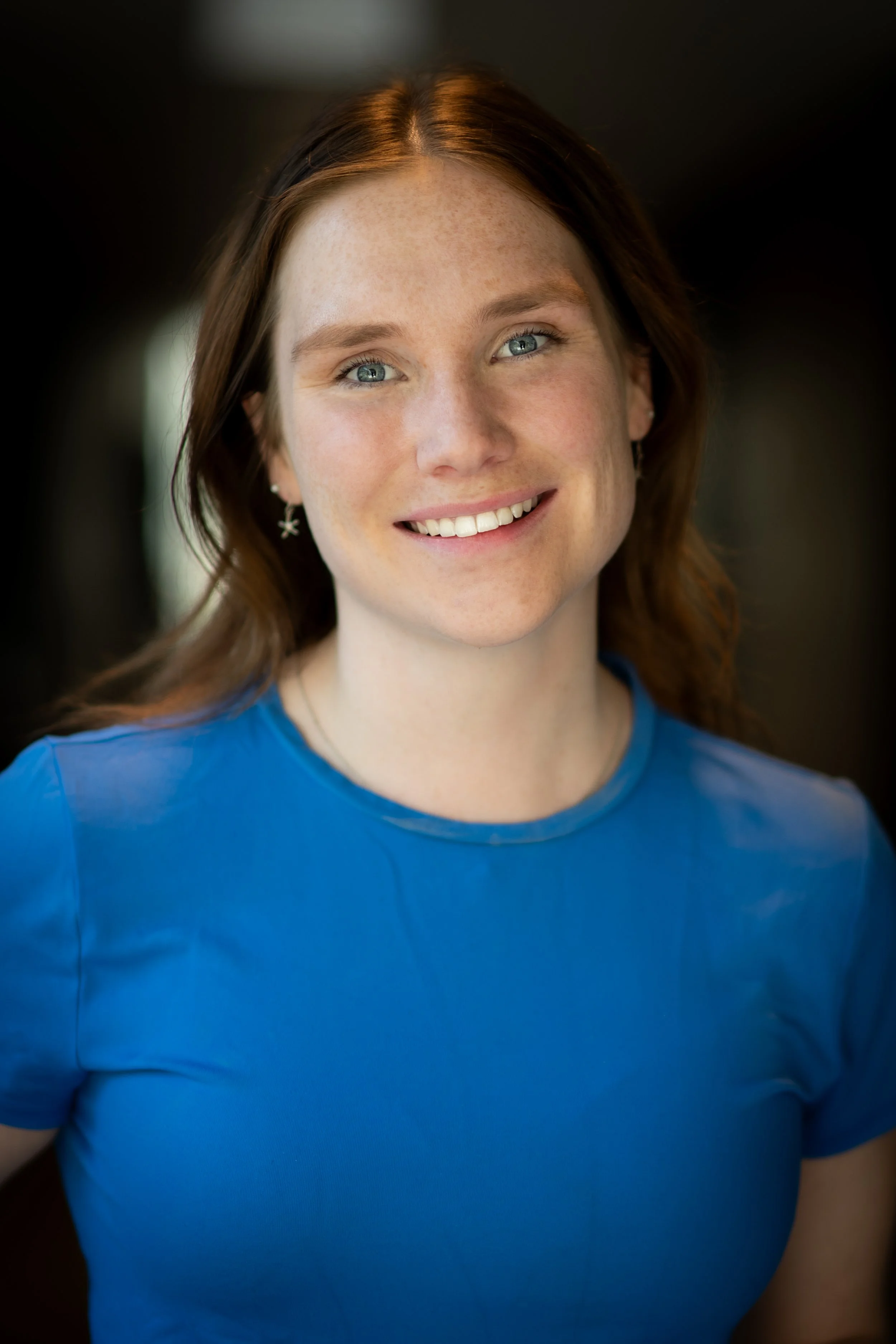 A young woman with red hair, blue eyes, and freckles, smiling in a blue shirt, looking at the camera.