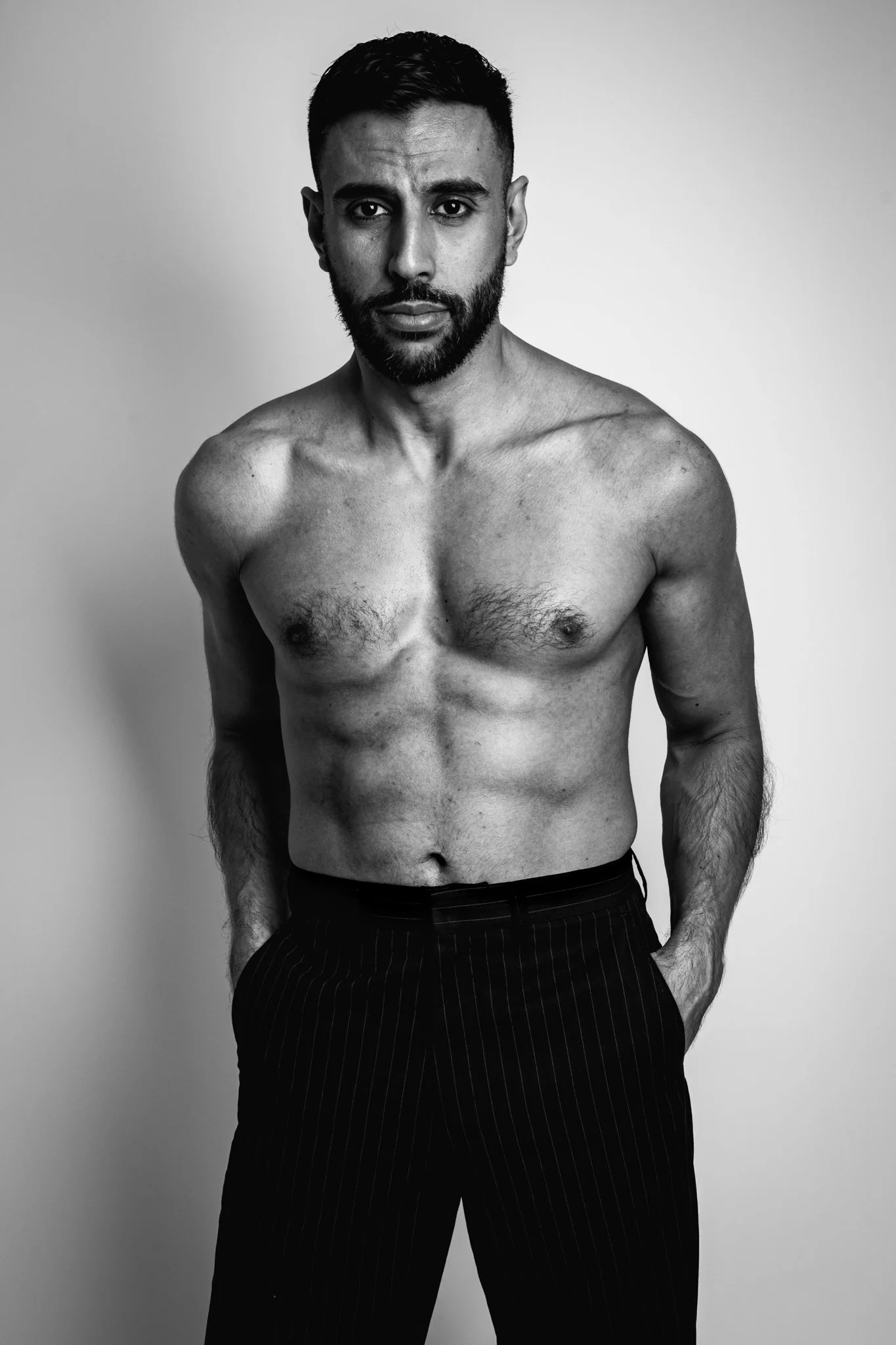 Black and white portrait of a shirtless man with a beard, looking directly at the camera, standing against a plain background.