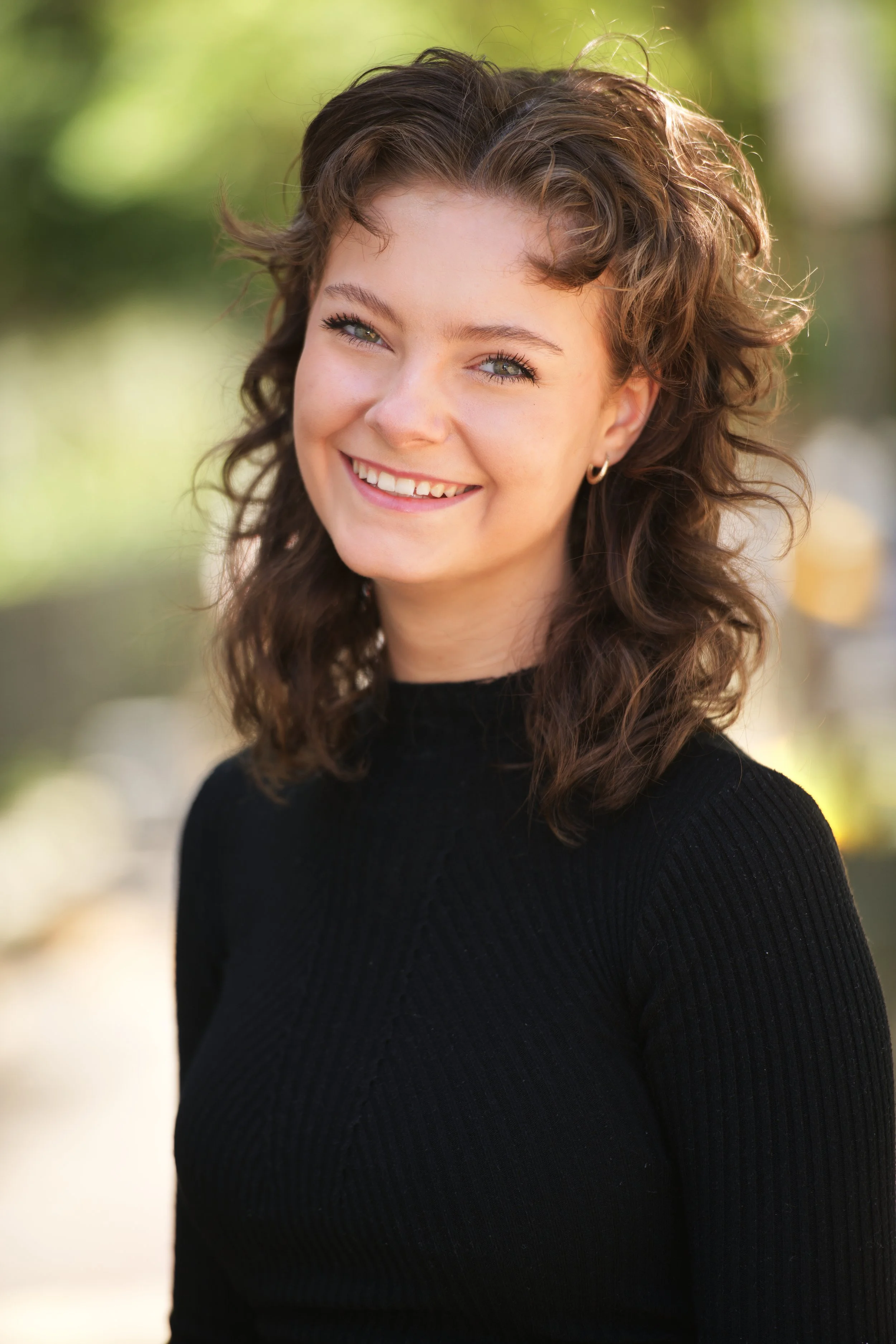 Young woman with curly brown hair, blue eyes, and small hoop earrings smiling outdoors with a blurred green background.