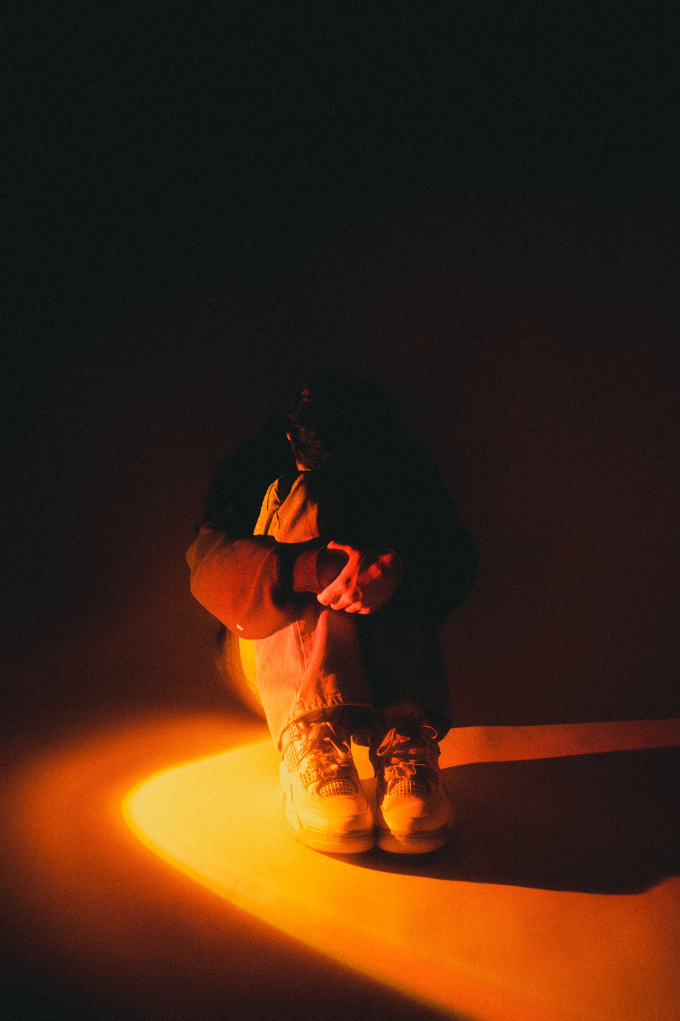 A person sitting on the ground in dim lighting with orange and yellow hues casting dramatic shadows.