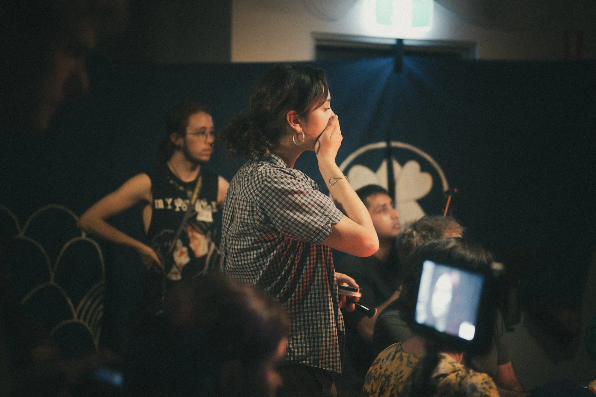 A woman with curly hair wearing a checkered shirt and hoop earrings, covering her nose with her hand, at an indoor event with a dark background and other seated people.