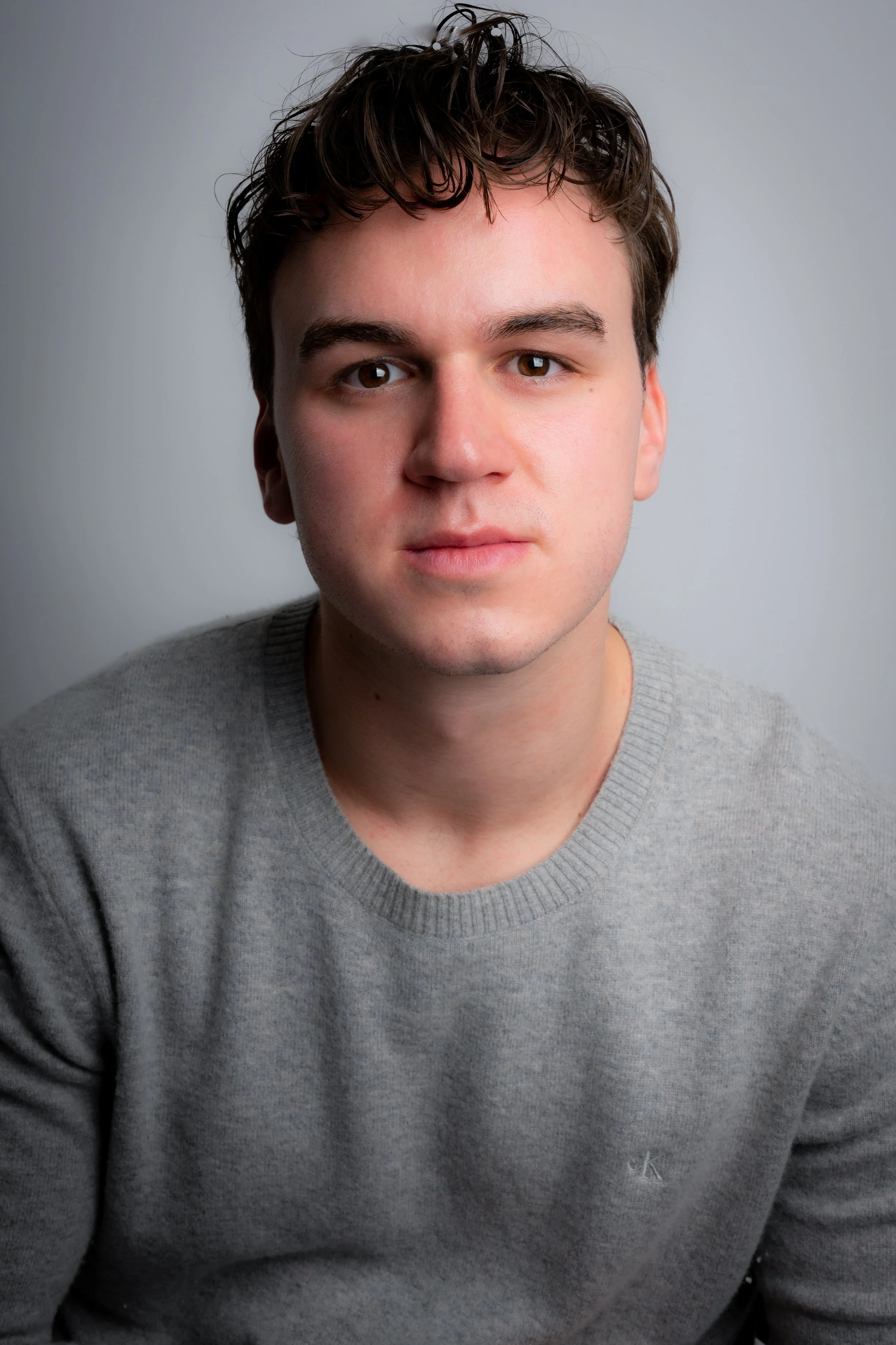 Portrait of a young man with wavy brown hair, wearing a light gray sweater, looking directly at the camera against a plain light background.