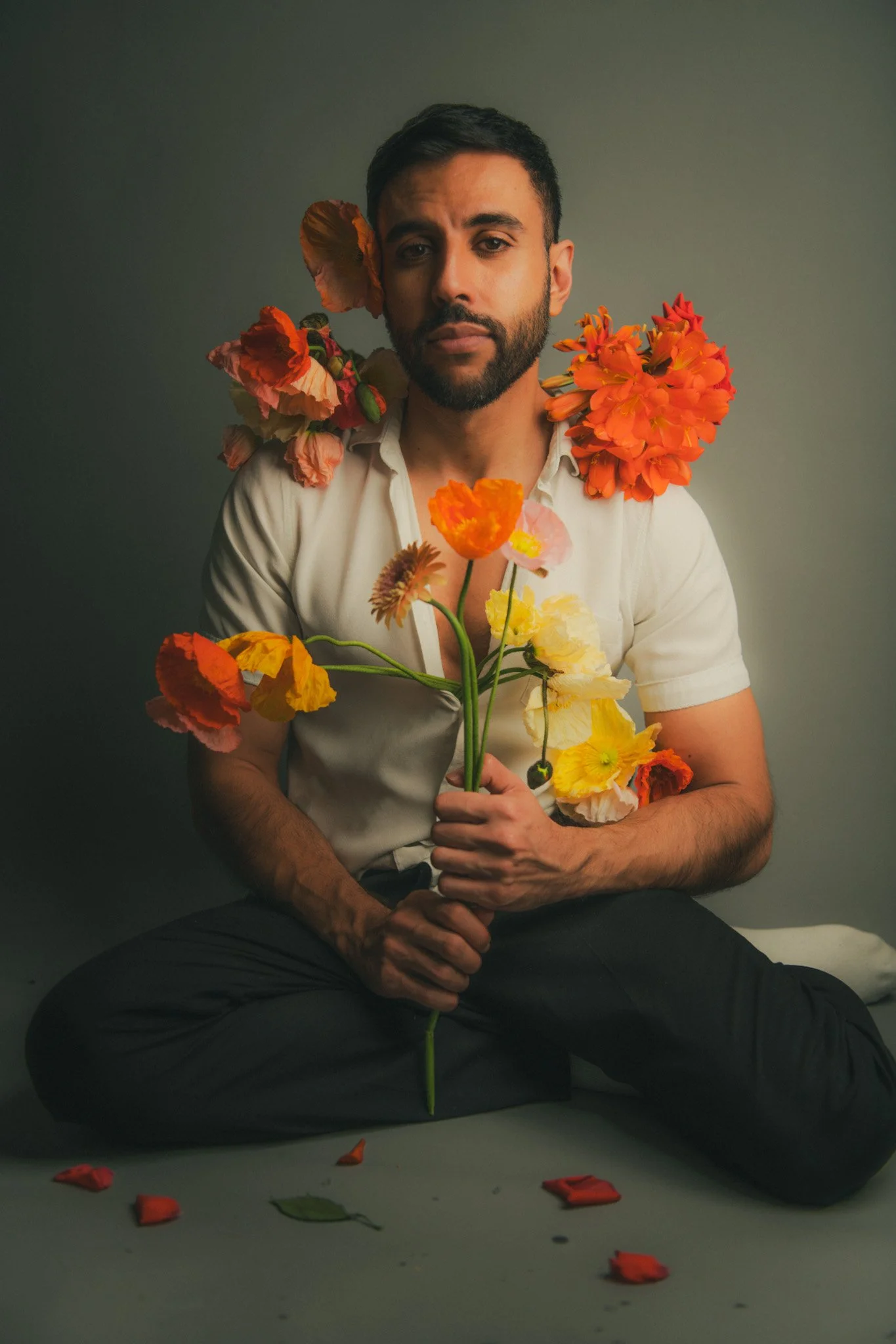 A man sitting cross-legged on the floor holding a bouquet of colorful flowers. He has a serious expression, with flowers around his neck and shoulders. Petals are scattered on the ground in front of him.