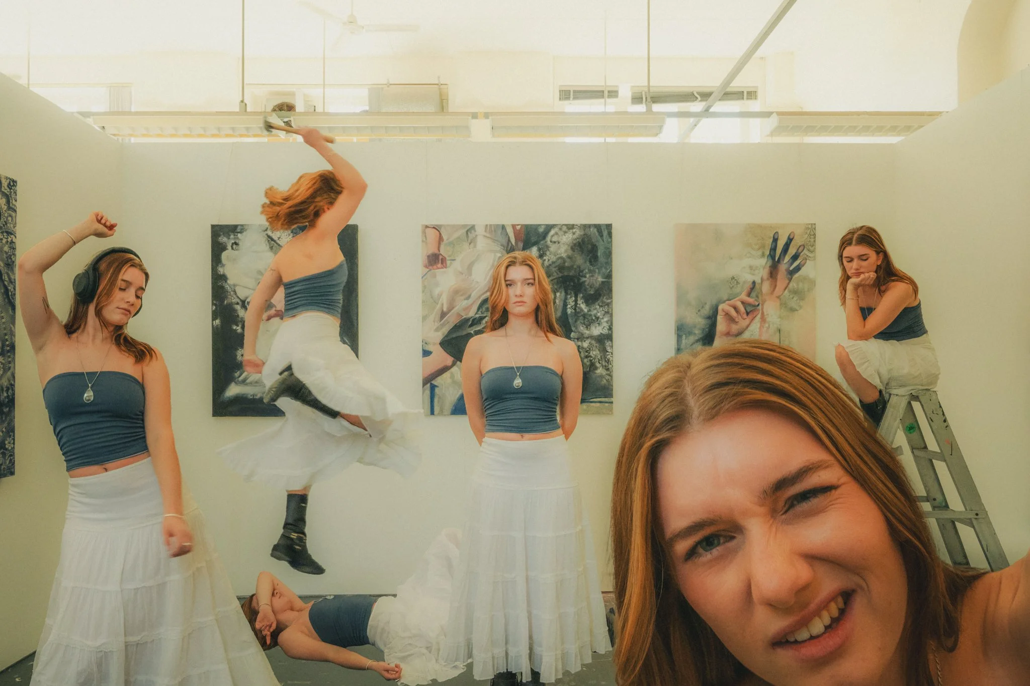 Several young women with red hair are posing in an art gallery. One is taking a selfie, another is on a ladder, and some are dancing or lying on the floor. They are dressed in matching tops and long white skirts, with paintings hanging behind them.