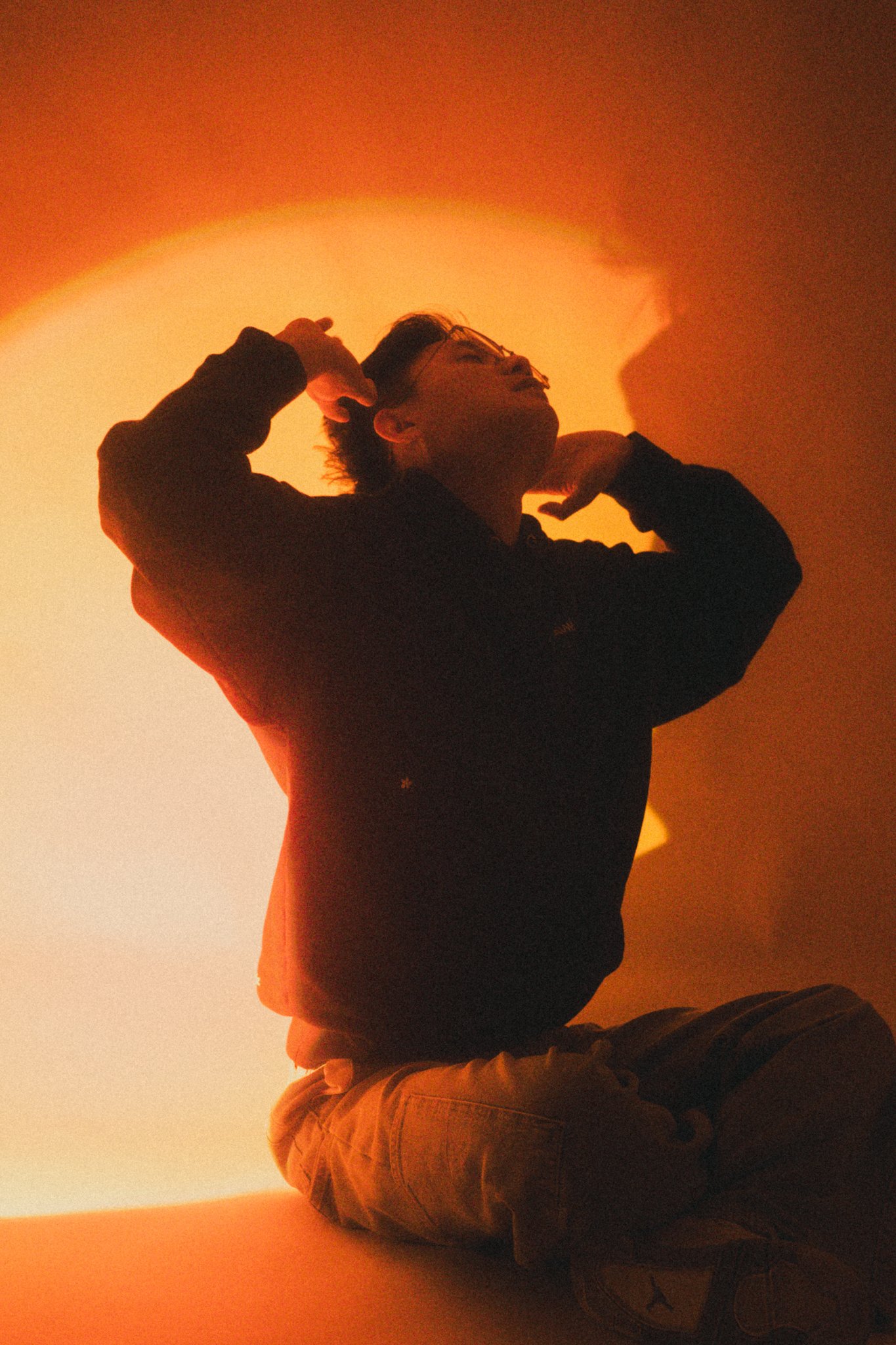 A person sitting cross-legged against a warm orange background, with an expressive pose and closed eyes.